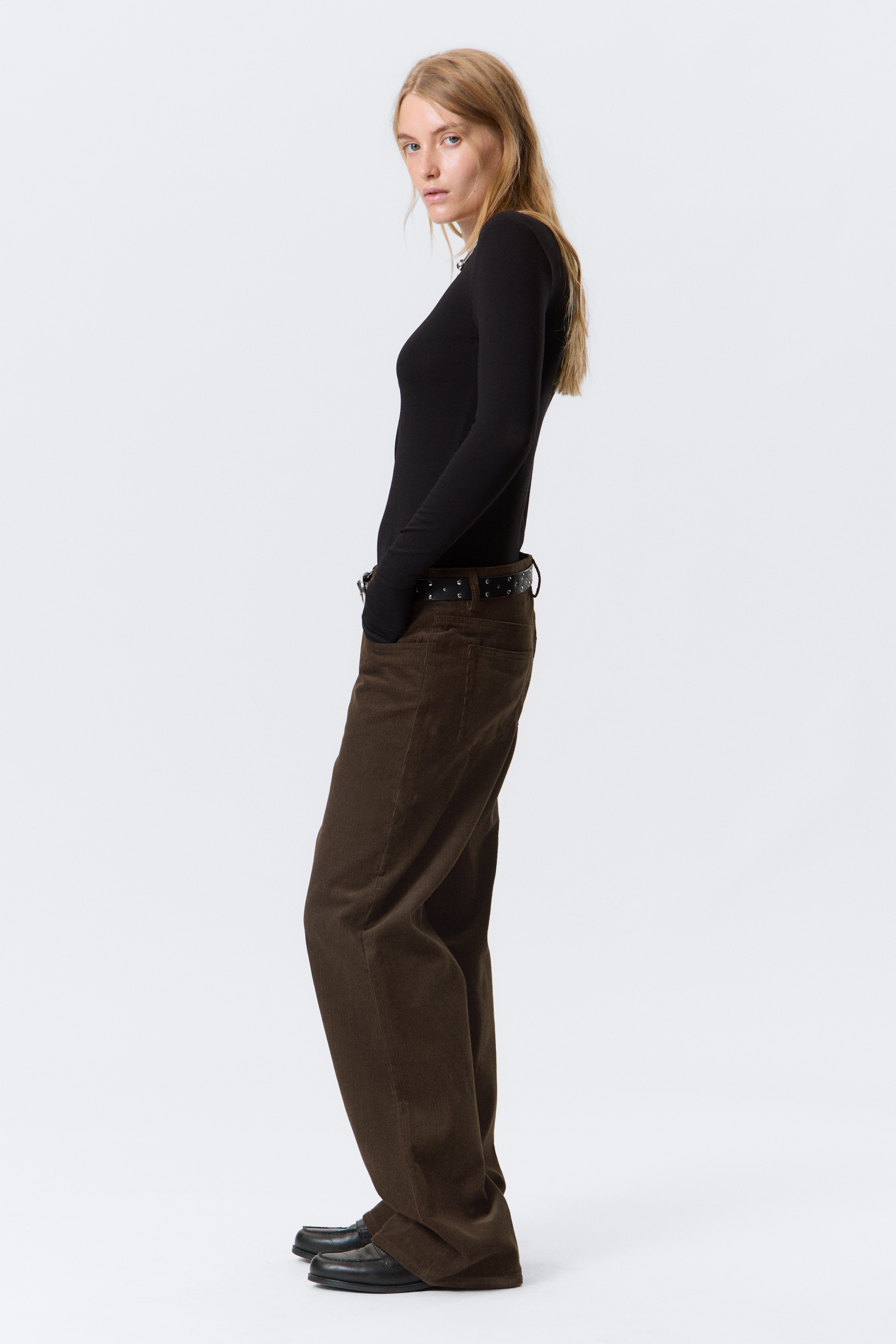 Visualizza immagine più grande: Visto da un'angolazione laterale, un top nero aderente a maniche lunghe con scollo rotondo è indossato infilato in pantaloni di velluto a coste marroni. I pantaloni presentano una vestibilità ampia e rilassata e sono abbinati a una cintura nera con borchie.