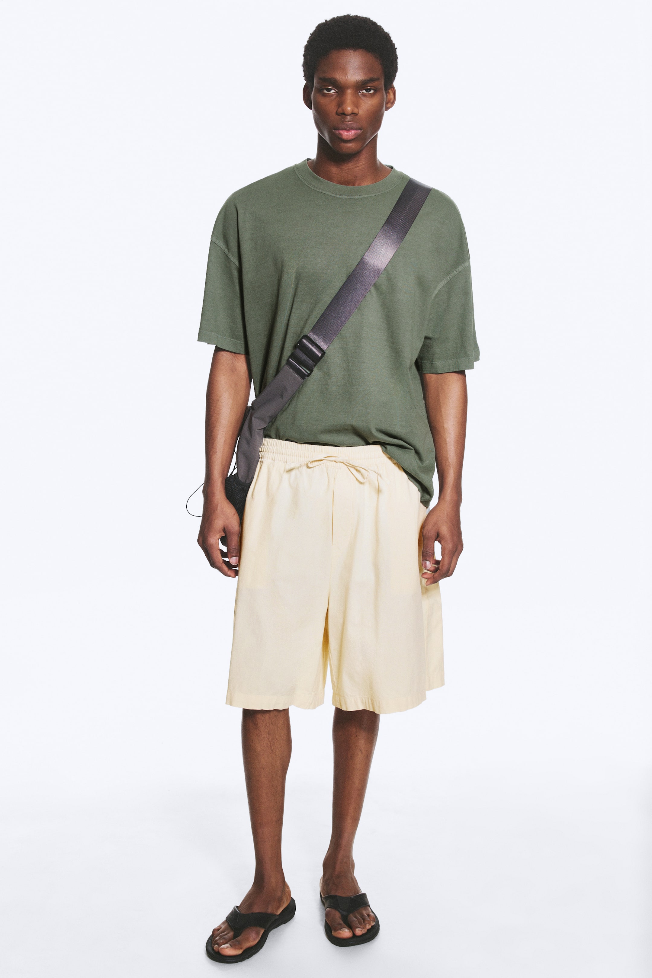 Größeres Bild anzeigen: Ein olivgrünes T-Shirt mit Rundhalsausschnitt, entspannter Passform und überschnittenen Schultern wird getragen, kombiniert mit cremefarbenen knielangen Shorts mit Kordelzug in der Taille. Schwarze Zehensandalen sind an den Füßen zu sehen, und eine dunkelgraue Umhängetasche mit breitem Riemen liegt quer über dem T-Shirt.