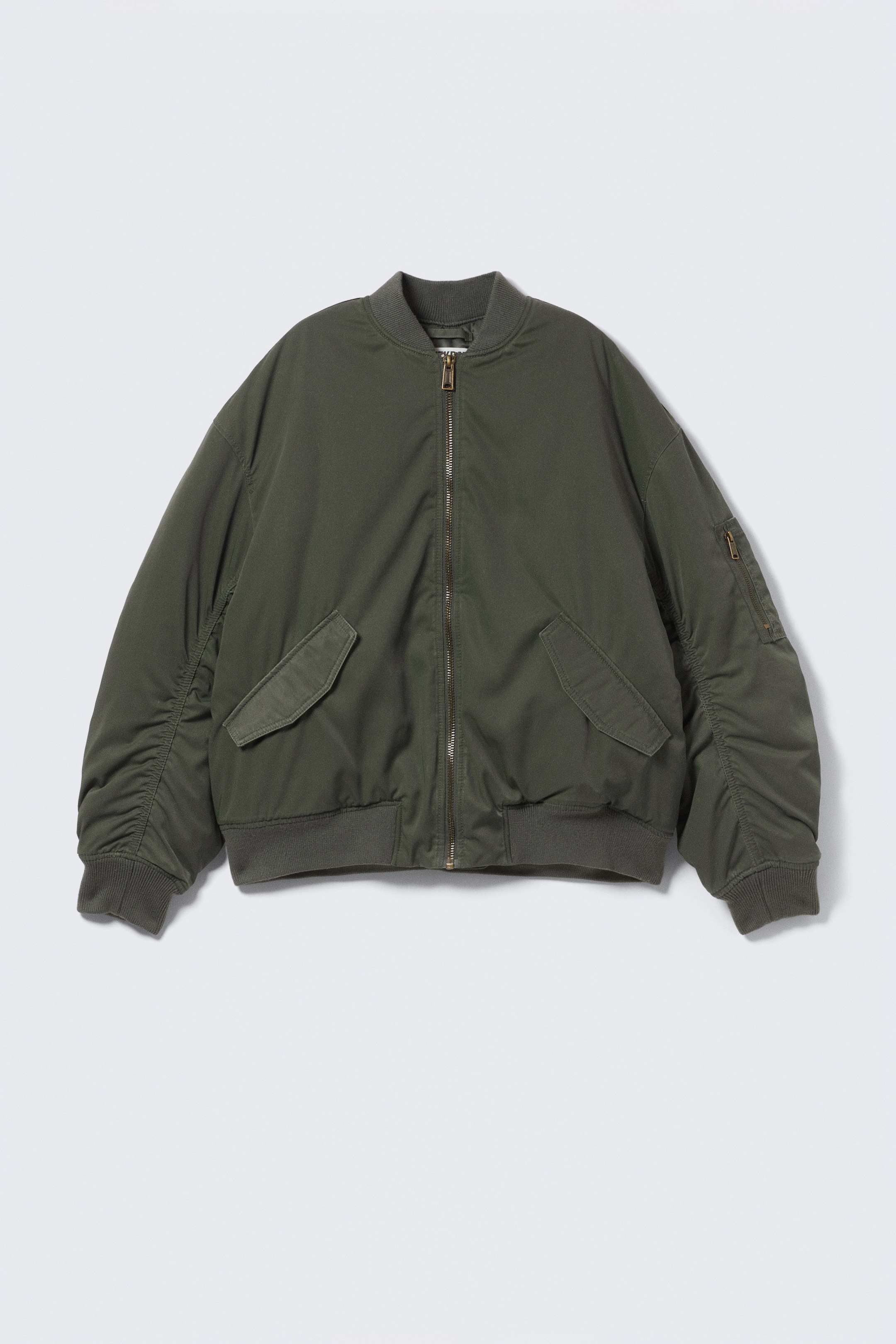 Visualizza immagine più grande: Una giacca bomber oversize verde oliva è presentata in PRIMA LINEA. Presenta una cerniera frontale centrale, due tasche con patta angolate sul davanti e una tasca con zip sulla manica sinistra. I polsini, il colletto e l'orlo a costine completano il design della giacca.
