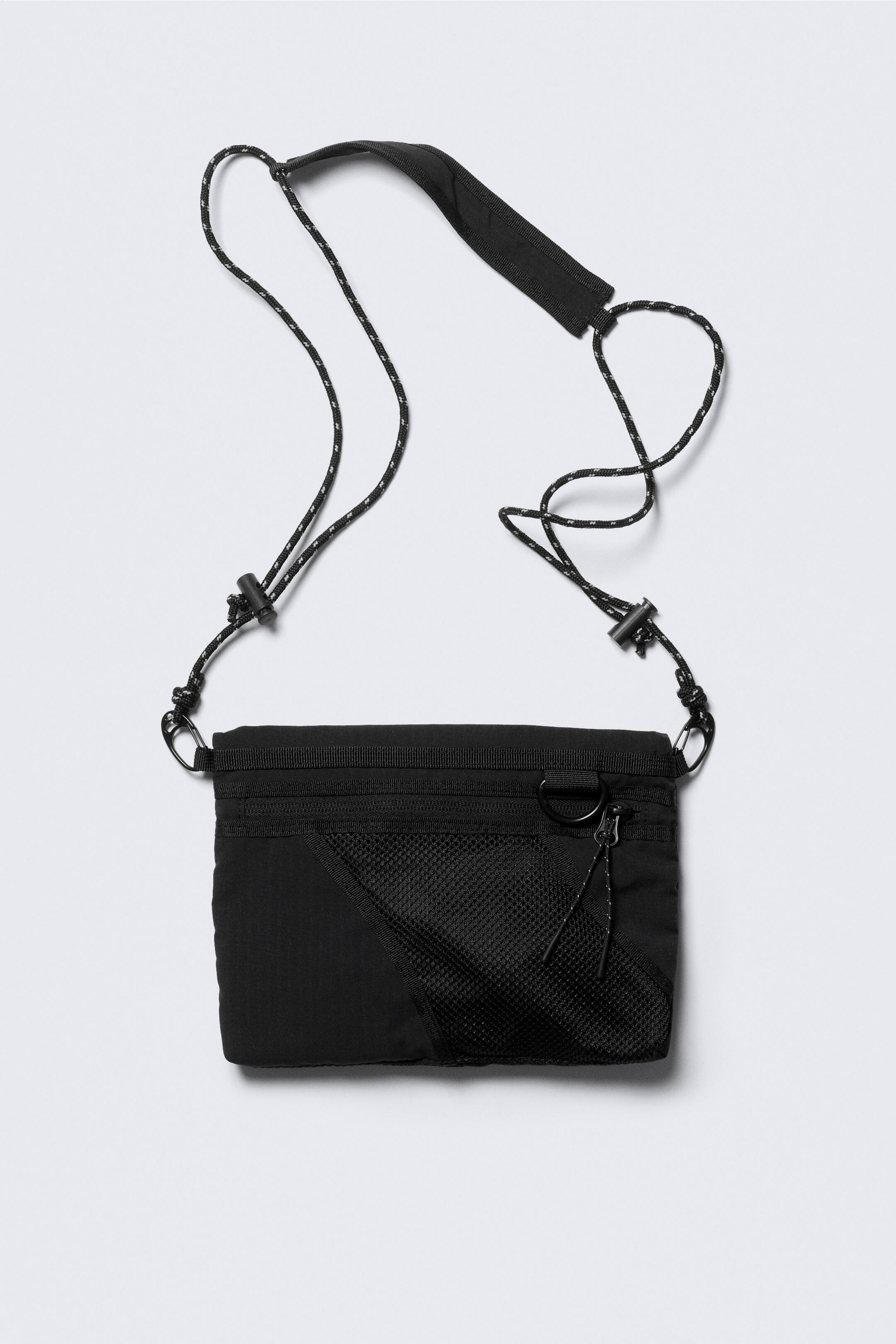 Vezi imaginea mai mare: O geantă neagră dreptunghiulară este afișată din față, având un buzunar cu fermoar orizontal și un buzunar din plasă diagonală cu fermoar și inel D în colțul din dreapta jos. O curea din cordon alb-negru cu secțiune căptușită și blocaje reglabile pentru cordon este atașată de geantă cu cleme carabinieră negre.
