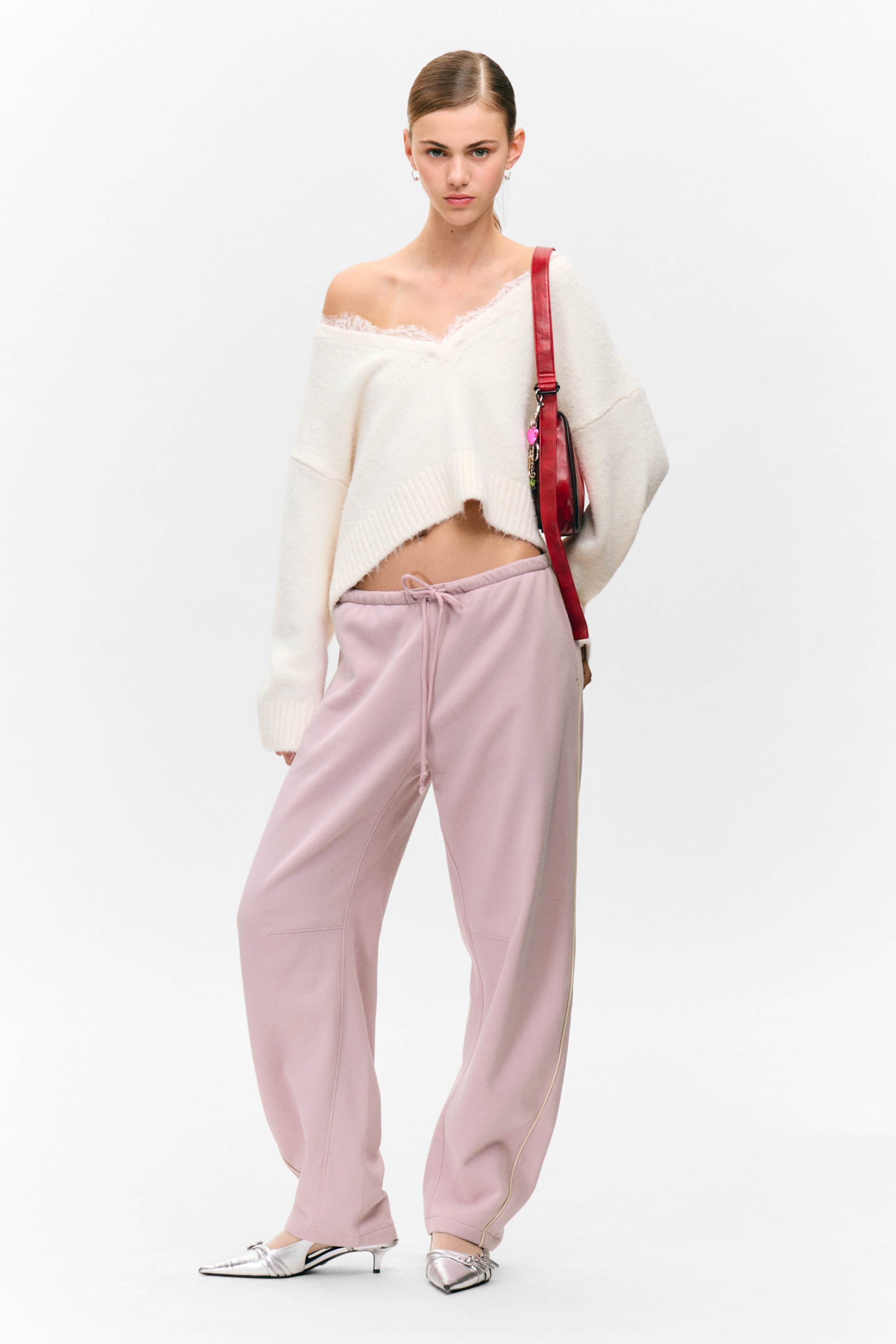 Ingrandisci l'immagine: Un maglione a V in maglia soffice color crema, con scollo rifinito in pizzo e orlo corto a coste, viene indossato con una spalla scoperta. È abbinato a pantaloni della tuta rosa chiaro a gamba larga, con vita con coulisse e una striscia verticale a contrasto su ciascun lato. Sono visibili anche dei tacchi a spillo argentati a punta e una borsa a tracolla rossa.