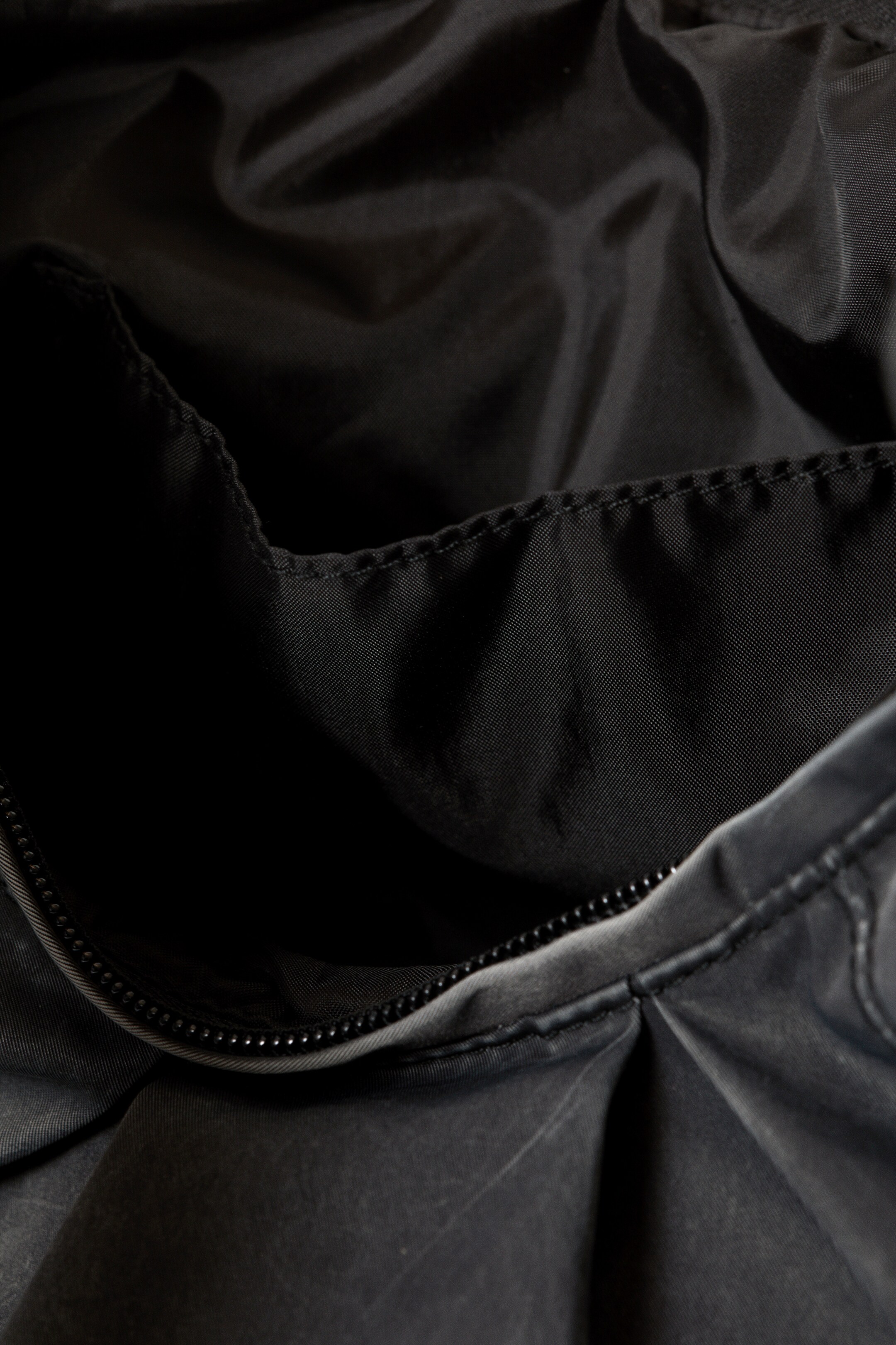 Grösseres Bild ansehen: Eine offene schwarze Tasche wird aus einer Vogelperspektive gezeigt, die ihren dunklen, strukturierten Außenstoff mit subtilen Falten und einem glatten, schwarzen Innenfutter offenbart. Ein schwarzer Reißverschluss ist entlang der Öffnung sichtbar und trennt die beiden Stoffe.