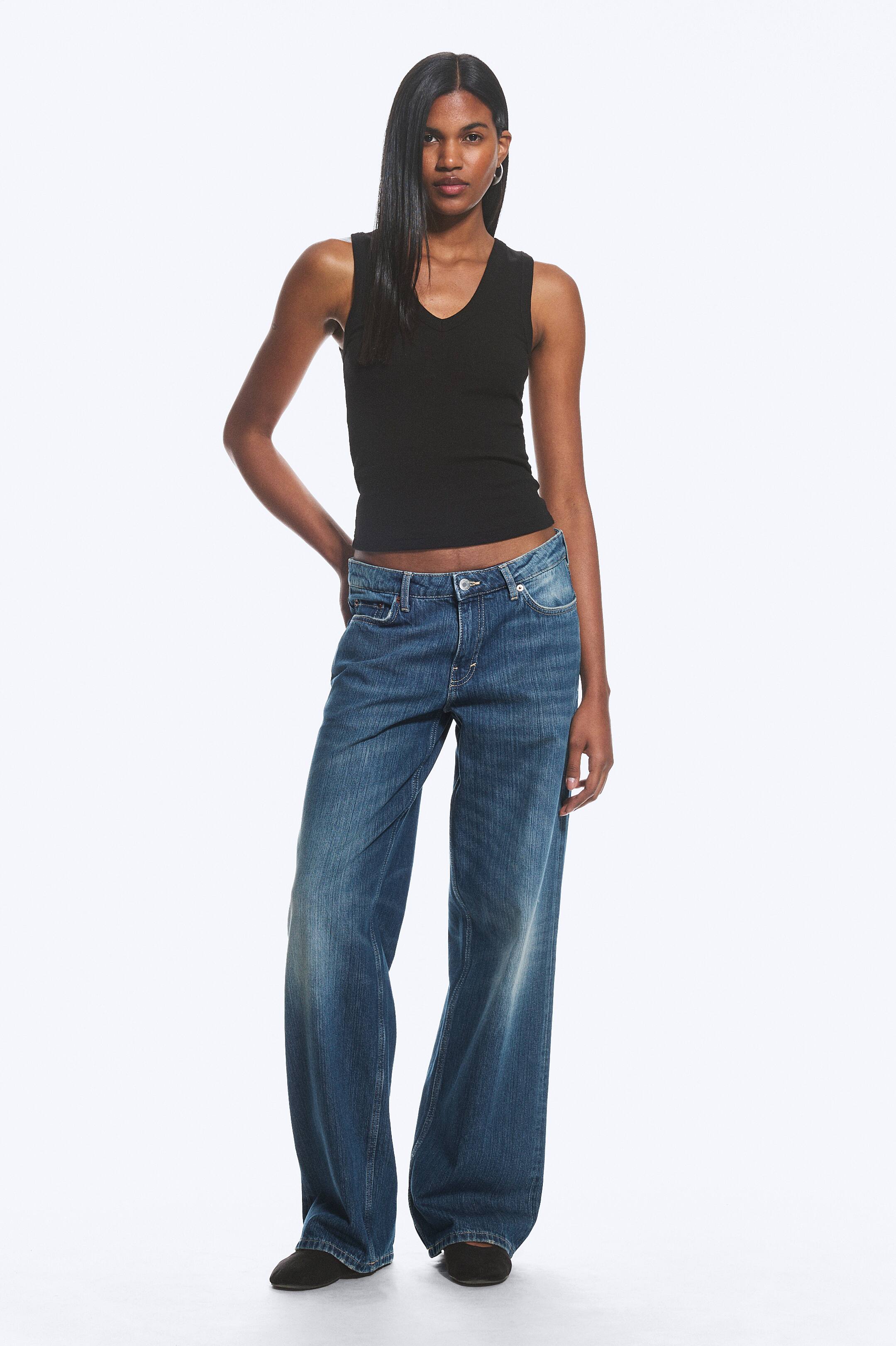 View larger image: A black ribbed V-neck tank top is being worn, showcasing a fitted silhouette. It is paired with blue wide-leg jeans, which feature a faded wash, a low-rise fit with a button and zip fastening, and relaxed legs gathering at the ankles.