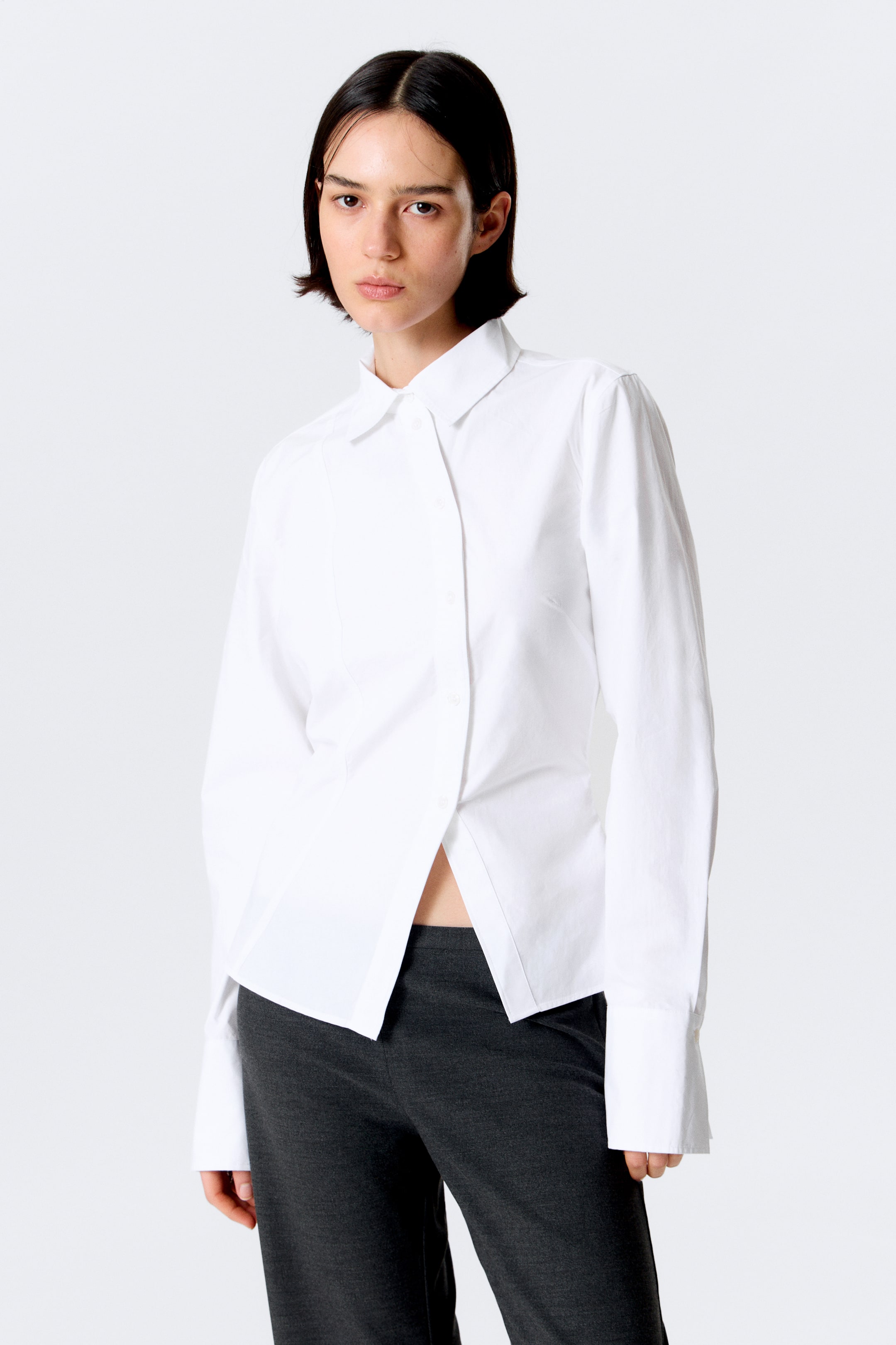 Ver imagen más grande: La modelo lleva puesta una camisa blanca de manga larga, que presenta un clásico cuello vuelto, una botonadura curva en el frente y una silueta entallada moldeada con pinzas visibles. Tiene un dobladillo asimétrico y puños anchos abotonados, y se combina con pantalones oscuros.