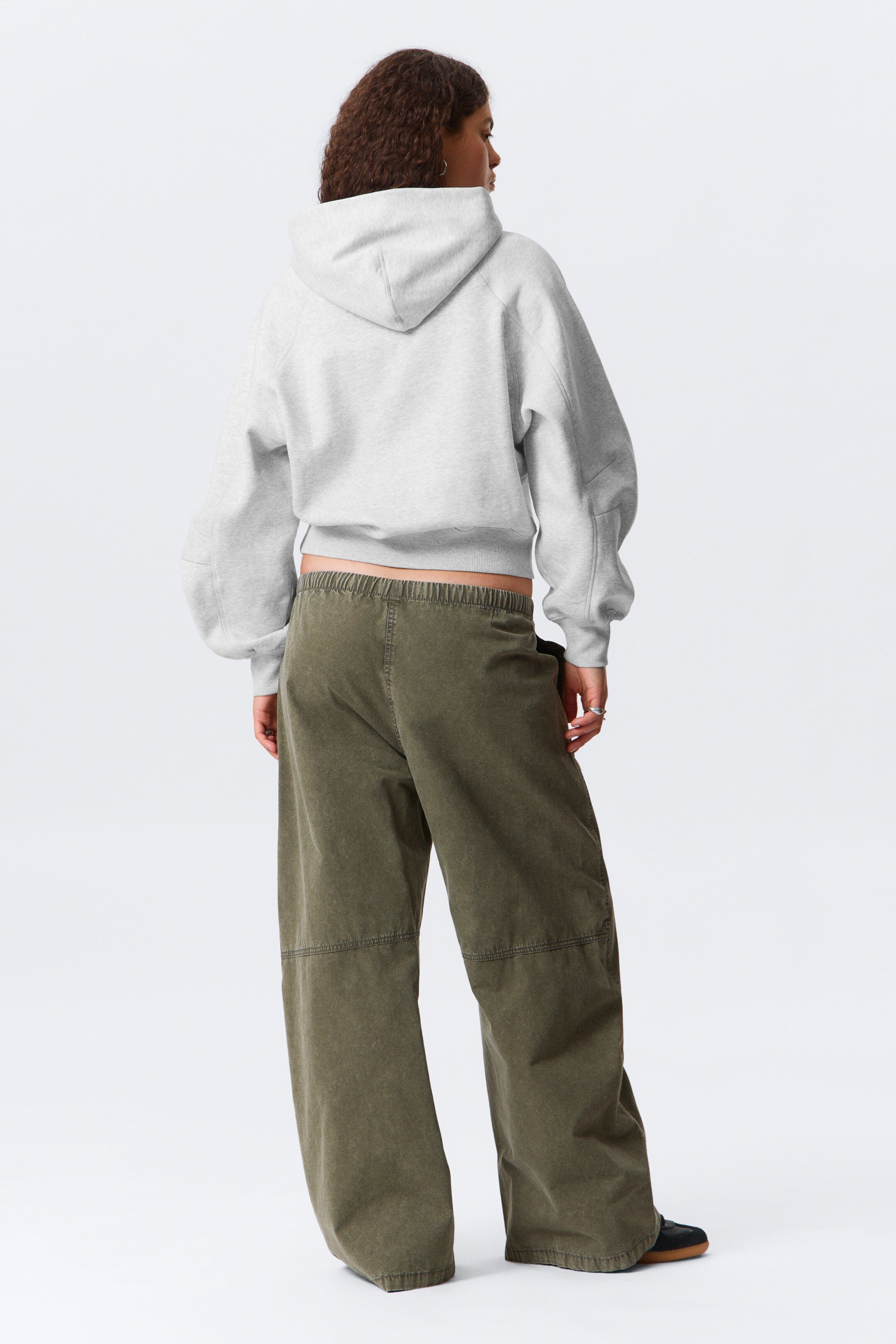 Visualizza immagine più grande: Da una vista posteriore, una felpa con cappuccio tagliata di colore grigio chiaro presenta un cappuccio e maniche raglan che si raccolgono ai polsini. Pantaloni a gamba larga di colore verde oliva mostrano un elastico in vita e dettagli delle cuciture lungo le gambe.