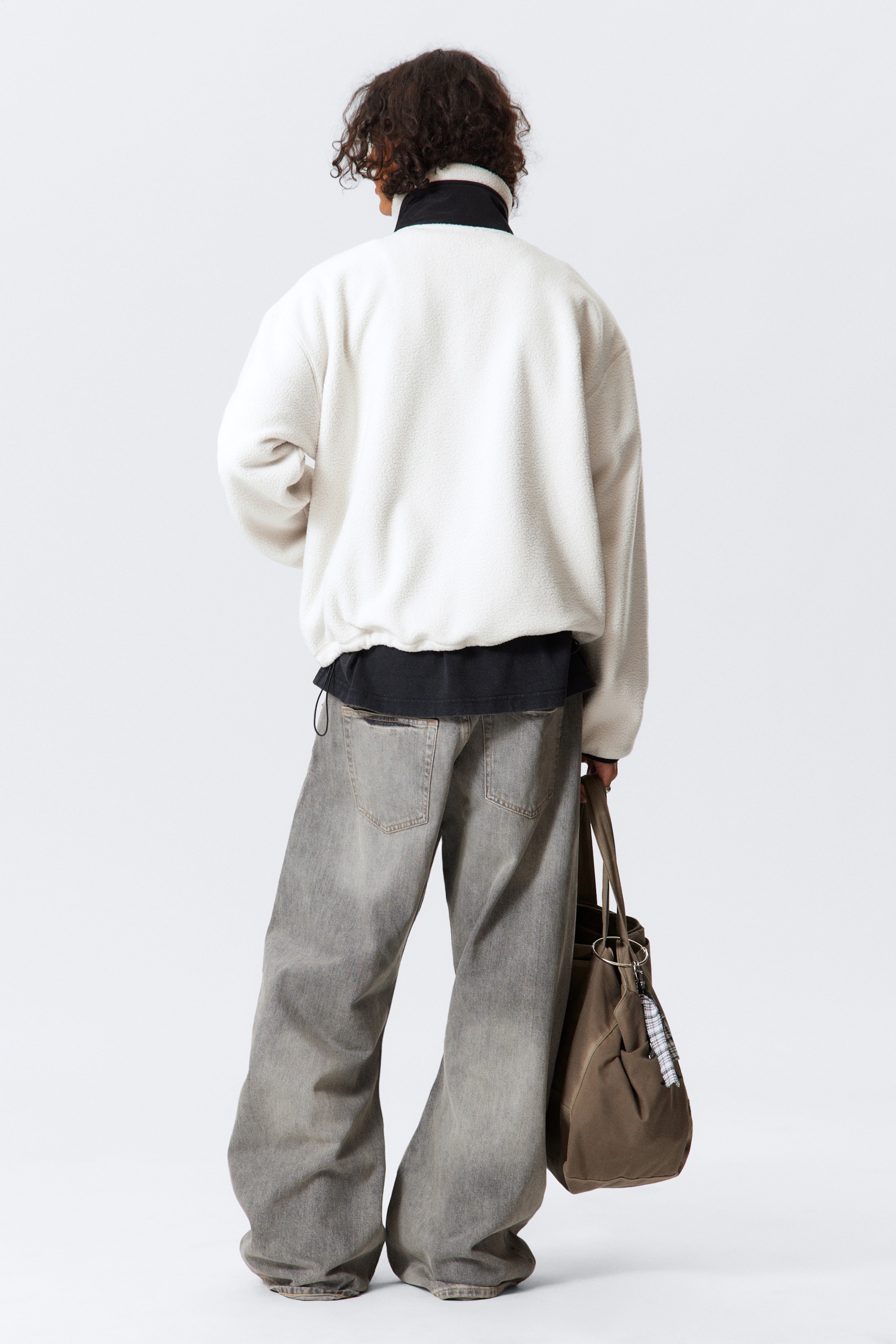 Visualizza immagine più grande: Una giacca in pile color crema a vestibilità rilassata è indossata, vista da dietro, con un colletto alto foderato di nero e un orlo con coulisse visibile. È stratificata sopra un top nero. Sono indossati anche jeans grigi sbiaditi a gamba larga con tasche posteriori visibili, e una borsa tote in tessuto marrone è tenuta in mano.