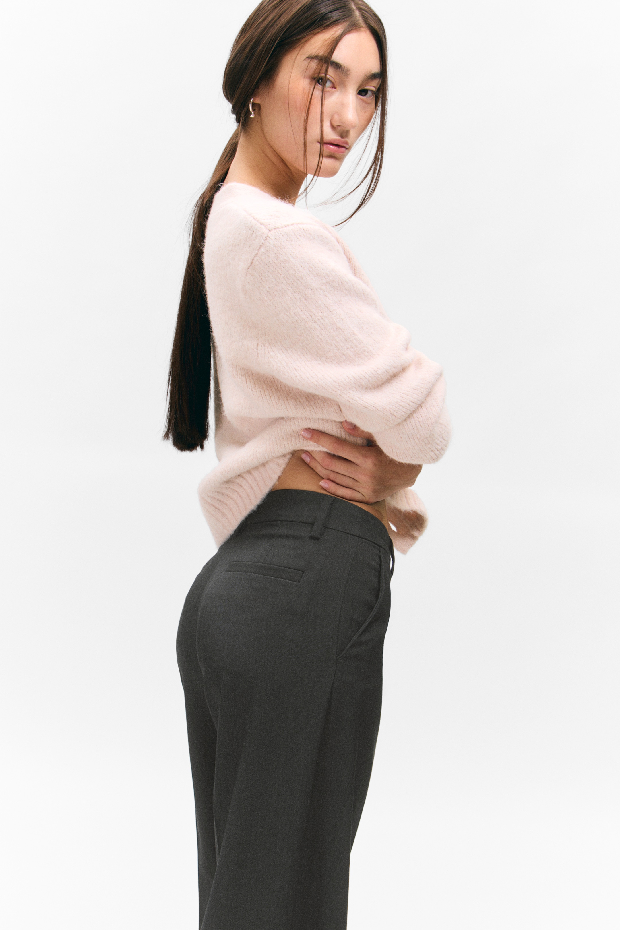 Größeres Bild ansehen: Ein hellrosa flauschiger Strickpullover wird getragen, der einen gerippten Saum hat, der hochgezogen wird. Dunkelgraue, maßgeschneiderte Hosen sind aus einem seitlich-hinteren Blickwinkel sichtbar und zeigen Gürtelschlaufen, Seitentaschen und eine einzelne hintere Paspeltasche.