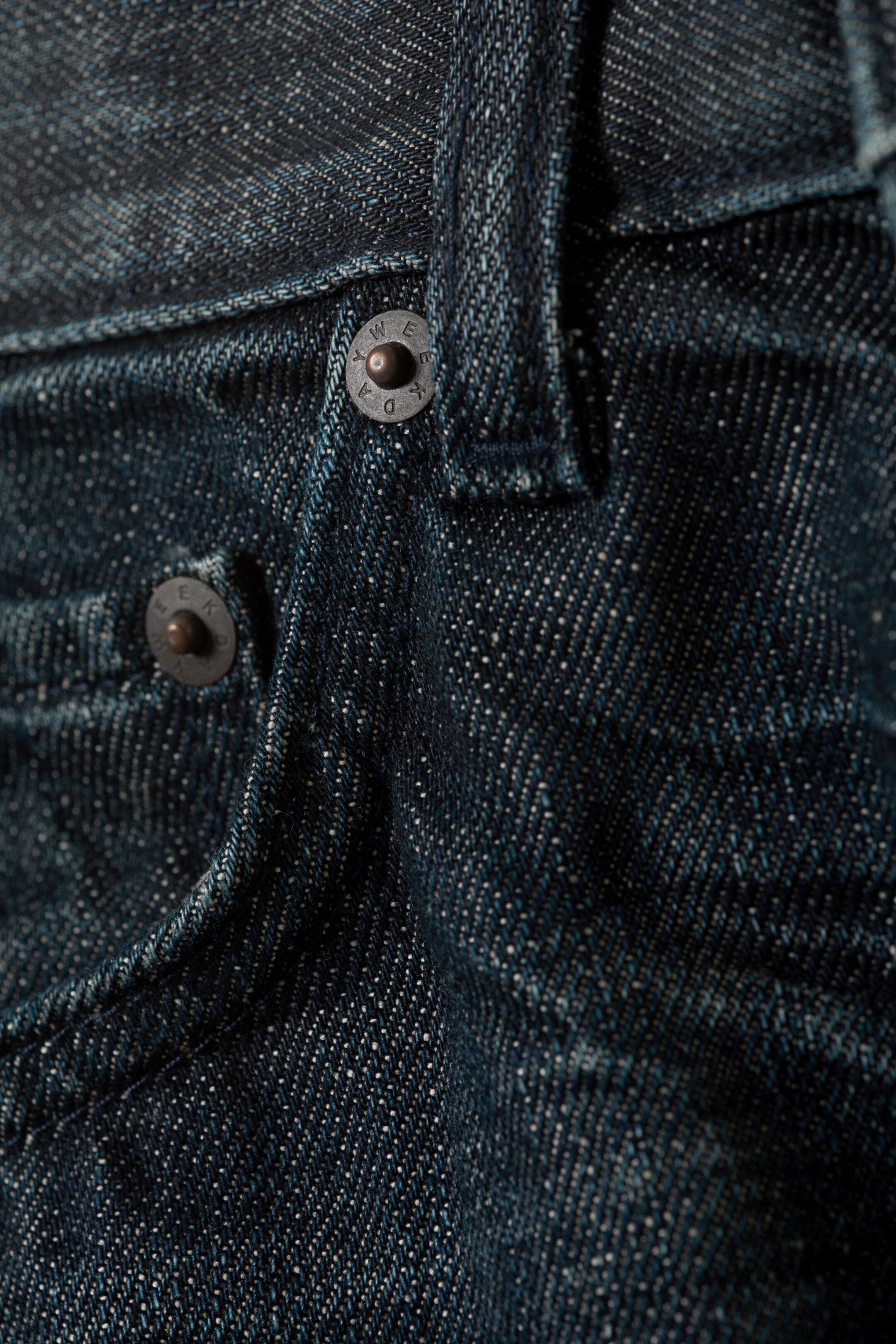 Se større billede: Mørkeblå denimjeans viser en nærbillede af deres teksturerede twillvævning. To mørke metalnitter, præget med tekst, sikrer stoffet nær en buet lommeåbning og en bæltestrop.