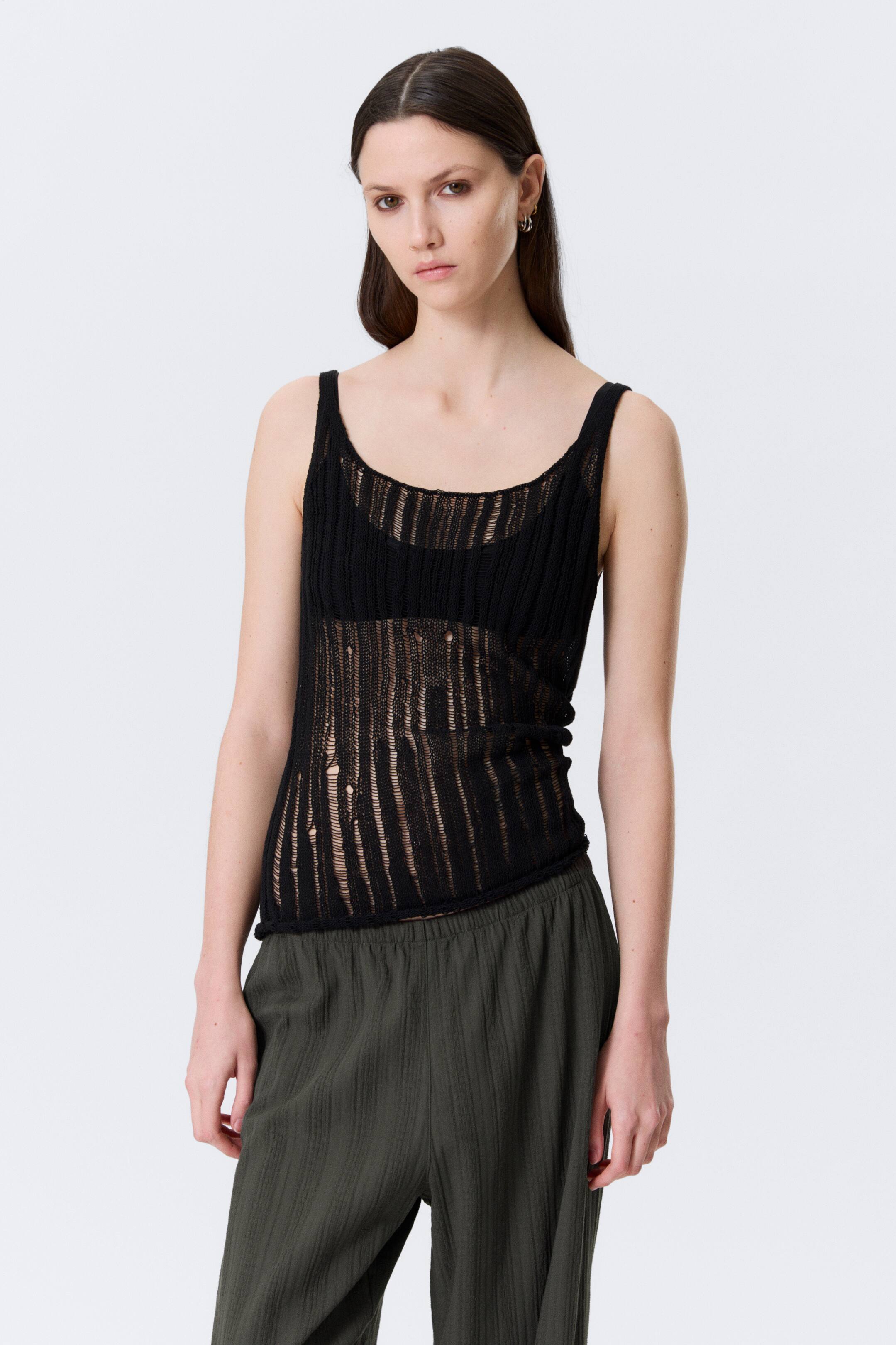 Visa större bild: En svart stickad linne med tunna axelband och en rund halsringning, visar ett slitet öppet stickmönster med vertikala trasiga områden över torson, bärs över en svart bh och vänd framåt.