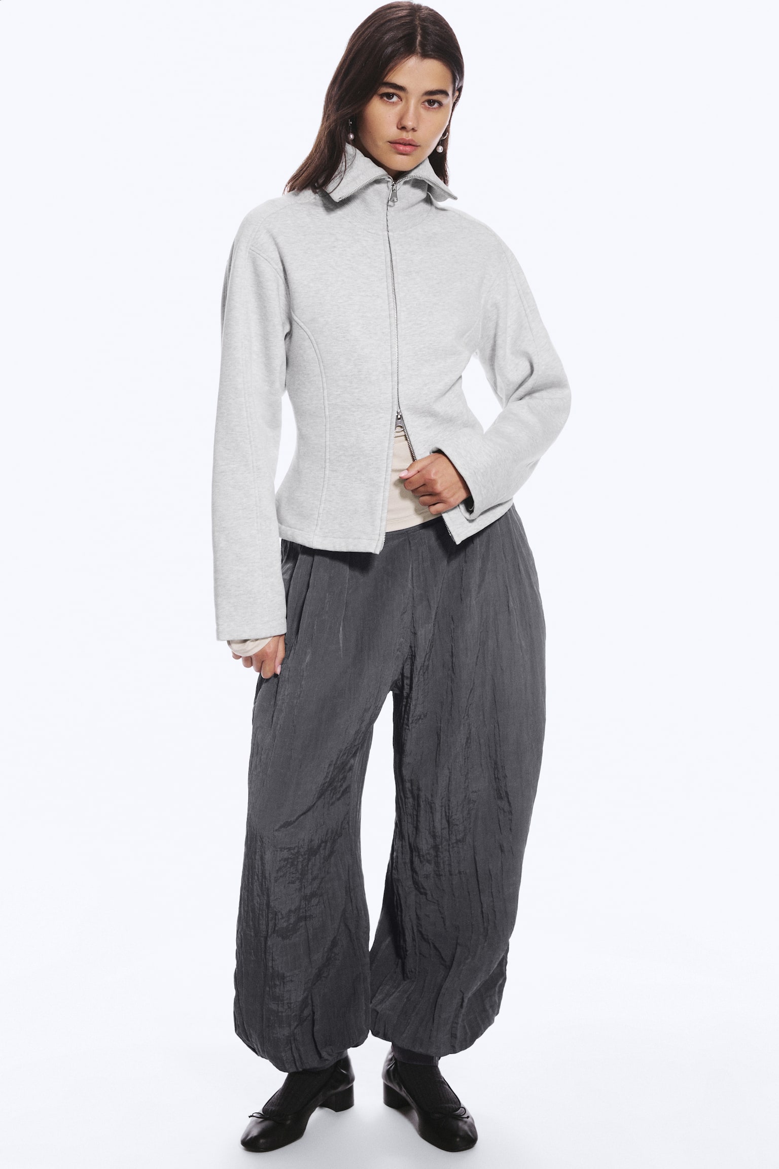 Una sudadera slim fit de color gris claro con cuello alto y costuras de panel curvadas la lleva la modelo, que sujeta el extremo inferior de la cremallera de doble dirección parcialmente abierta. La sudadera se combina con una prenda beige y se lleva con pantalones anchos de textura en gris oscuro. - 1
