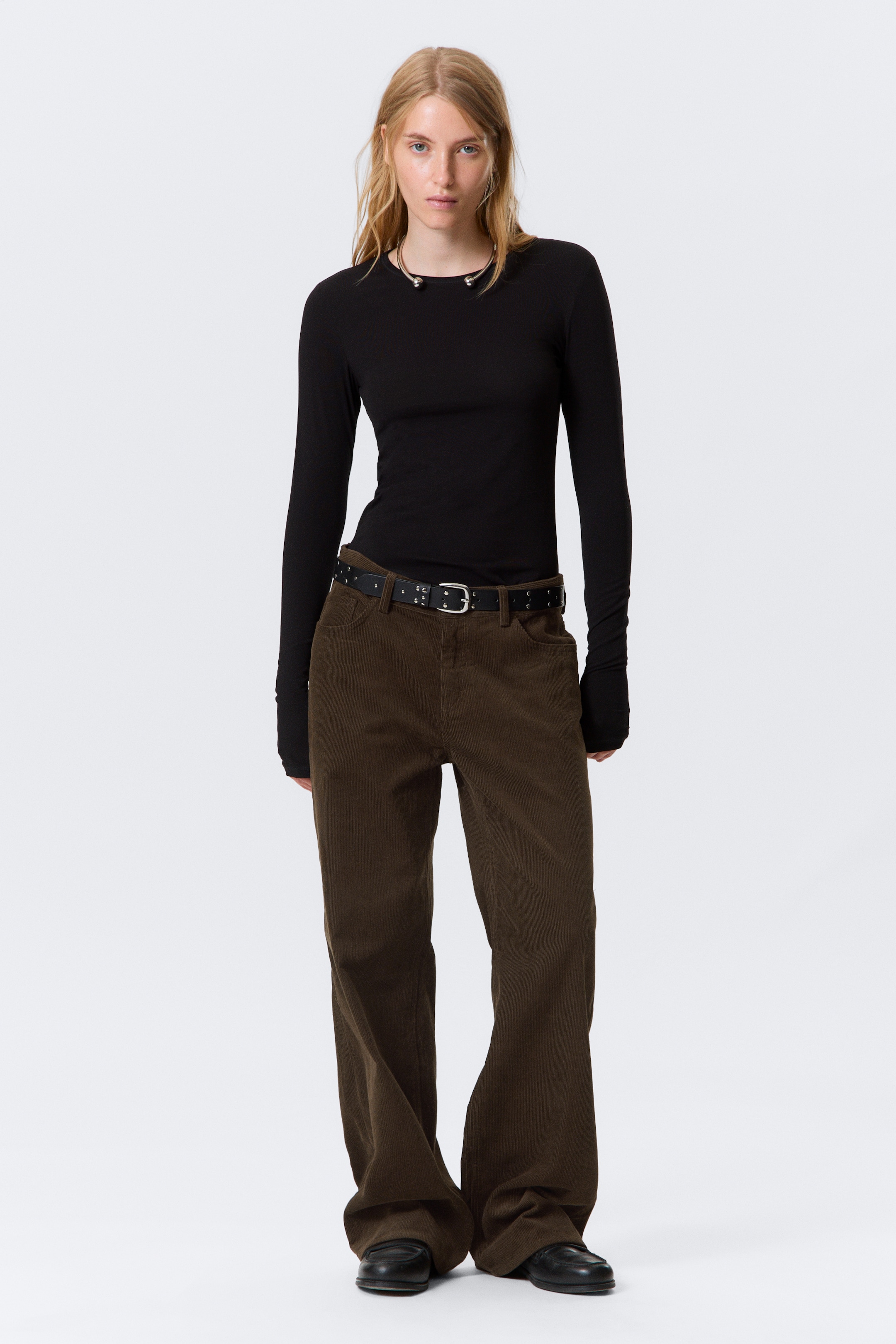 Visualizza immagine più grande: Una persona indossa un top nero aderente a maniche lunghe con fori per i pollici e pantaloni a gamba larga in velluto a coste marrone scuro. I pantaloni presentano tasche frontali visibili e sono abbinati a una cintura nera con borchie, fissata da una fibbia rettangolare color argento.
