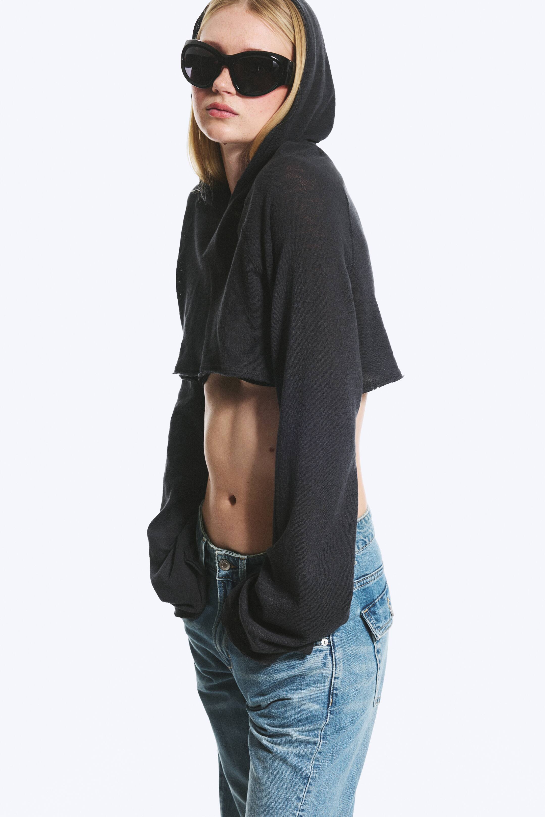 Agrandir l'image: Un hoodie court noir est présenté en vue légèrement de côté, avec une capuche et des manches longues et amples dotées de poignets à coupe brute. Il affiche une ligne d'ourlet à coupe brute qui s'élève au-dessus de la taille, et son tissu léger crée une coupe décontractée.
