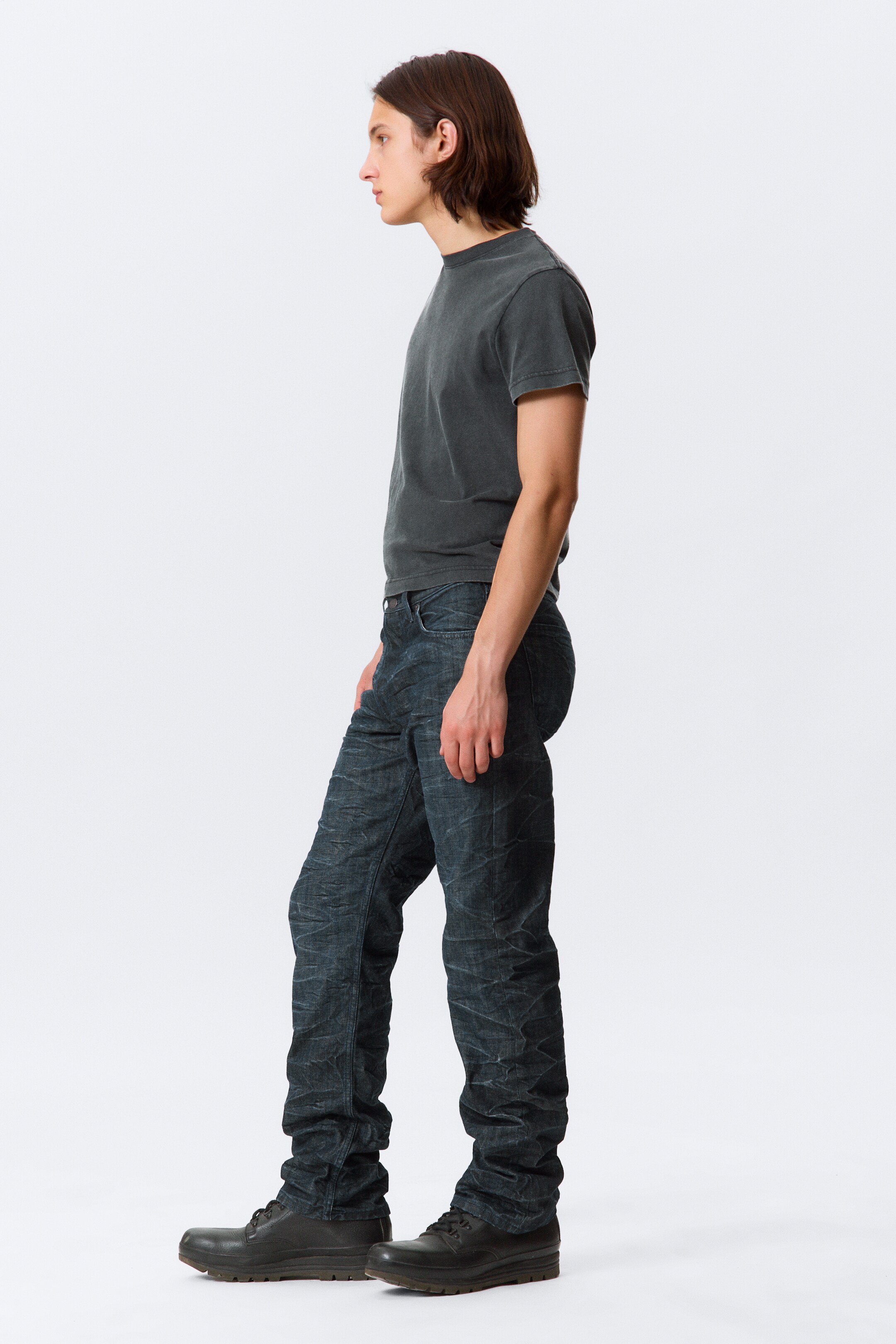 Se større billede: En mørkegrå, afslappet pasform, kortærmet t-shirt med rund hals bæres sammen med mørke jeans, der har et markant krøllet og tekstureret udseende og samler sig ved anklerne over mørke snøresko med tykke, riflede såler.