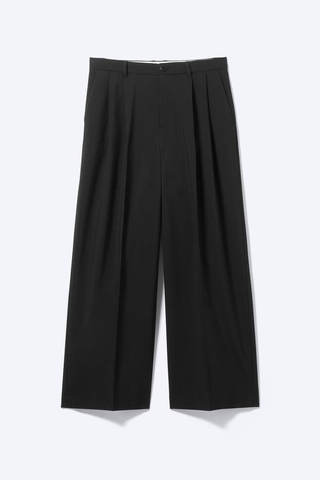 Ein Paar schwarze Twill-Anzughosen wird von vorne präsentiert und zeichnet sich durch eine lockere, weite Passform aus. Sie sind mit einem Reißverschluss und einem Knopf am Bund ausgestattet, haben schräge Seitentaschen, vordere Falten und gebügelte Bügelfalten entlang der Vorderseite jedes Beins. - 1
