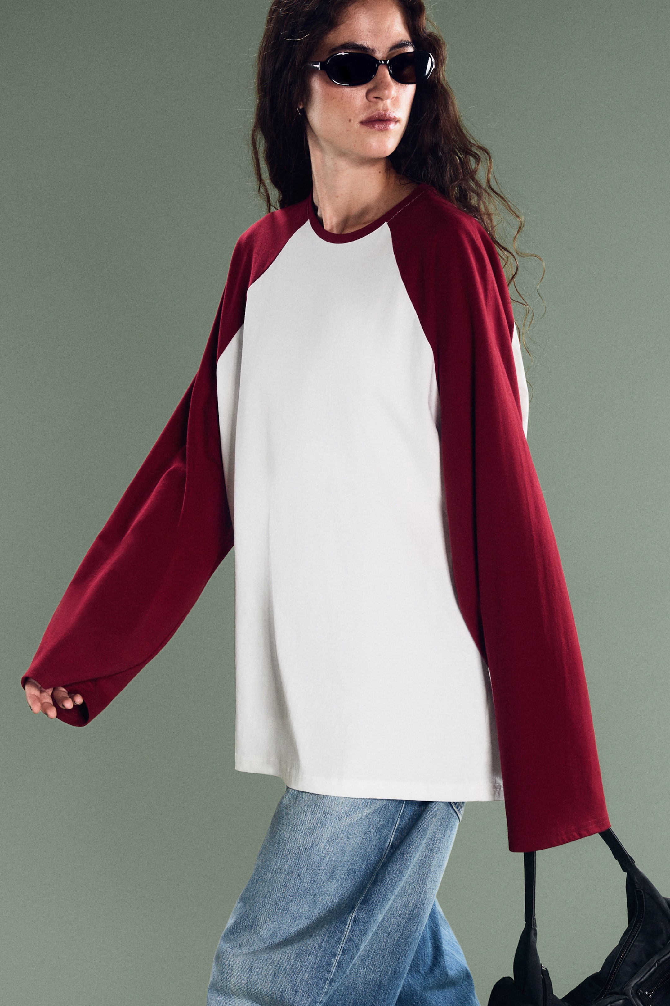 Zobacz większy obraz: Model ma na sobie luźną bluzkę z długim rękawem typu raglan, z białym korpusem i bordowymi rękawami. Oversize’owy model ma okrągły dekolt oraz szerokie, bardzo długie rękawy i został zestawiony z jasnoniebieskimi jeansami.