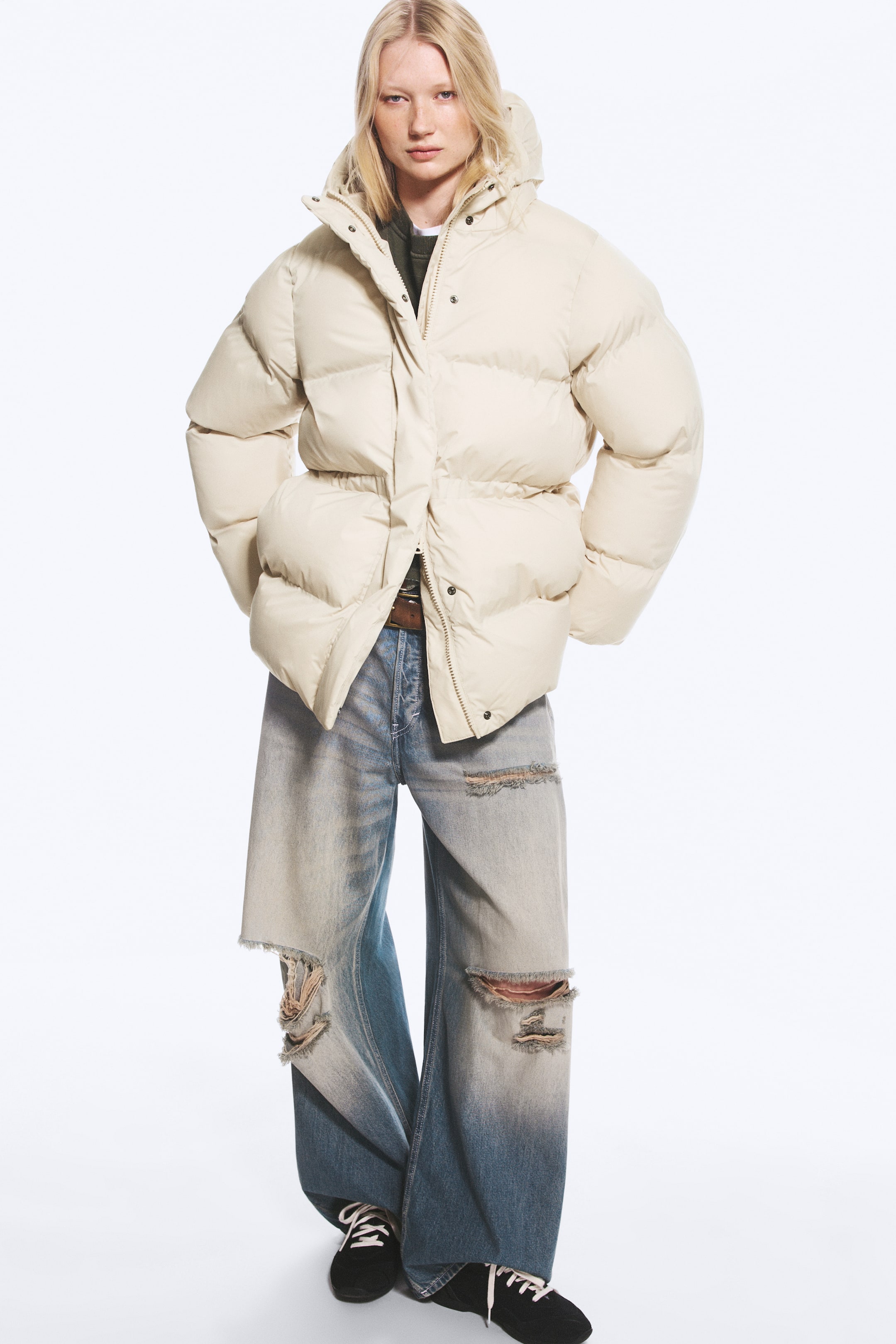 View larger image: A cream-coloured waisted puffer jacket featuring horizontal quilting, a hood, and a gathered waist is being worn by the model. The jacket is styled partially open, layered over a dark top and paired with distressed, wide-leg blue jeans.