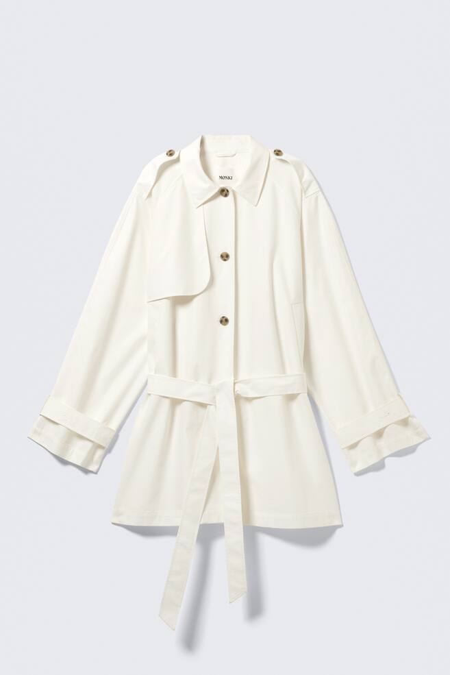 A short, single-breasted trench coat in an off-white colour is being displayed from the FRONT, featuring a loose fit, a turn-down collar, buttoned epaulettes, and a gun flap on the left chest. A tie belt is cinching the waist, and the long sleeves are ending with adjustable cuffs that are rolled up. - 1