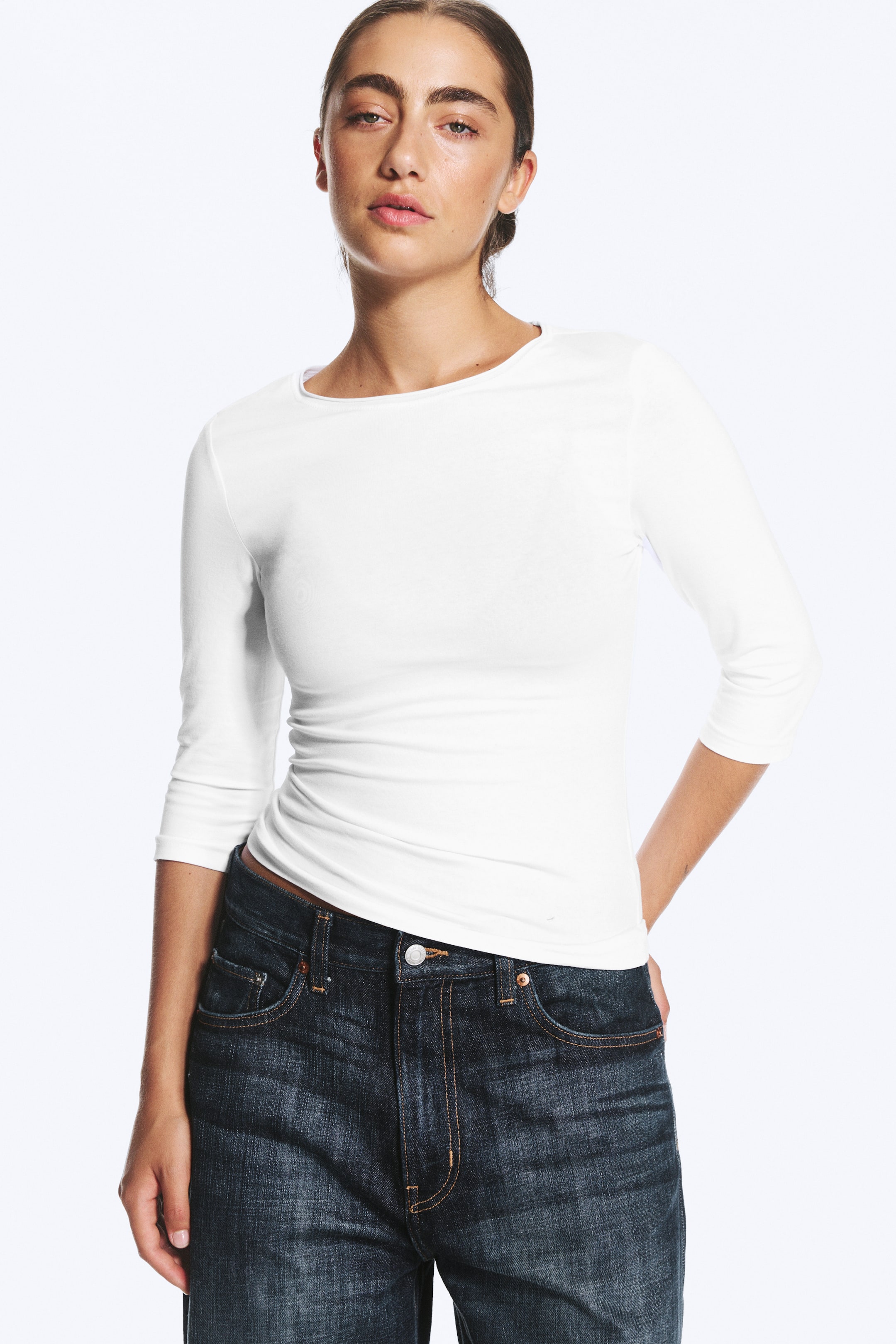 Grösseres Bild ansehen: Das Model trägt ein weisses, eng anliegendes Oberteil mit Rundhalsausschnitt und Dreiviertelärmeln, kombiniert mit dunkelblauen Jeans.