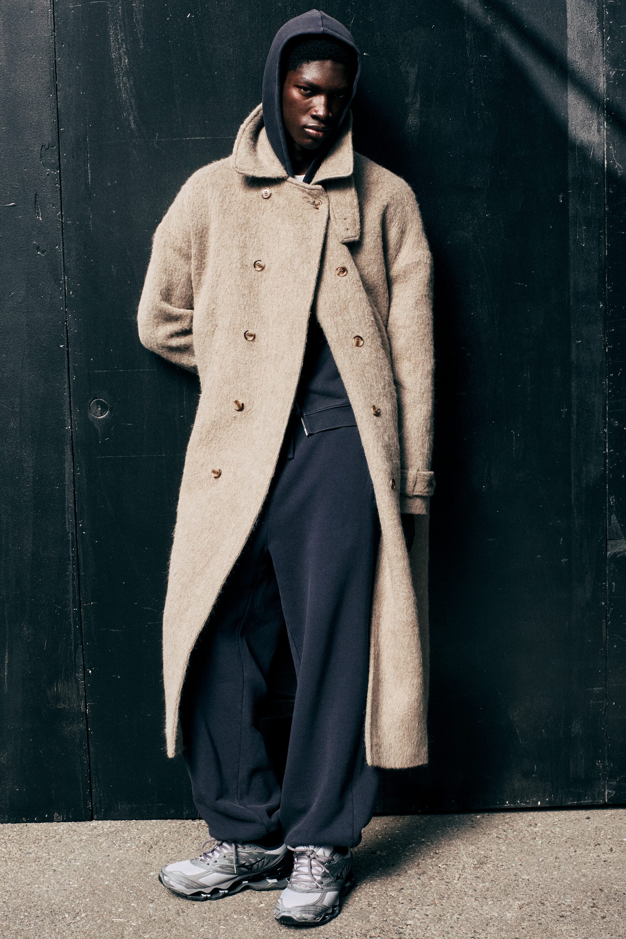 A long, oversized wool-blend coat in a textured beige shade is shown from the front, featuring a double-breasted button fastening and a high, buttoned stand collar. The coat is styled with slanted jetted pockets and adjustable tabs at the cuffs of its relaxed sleeves.