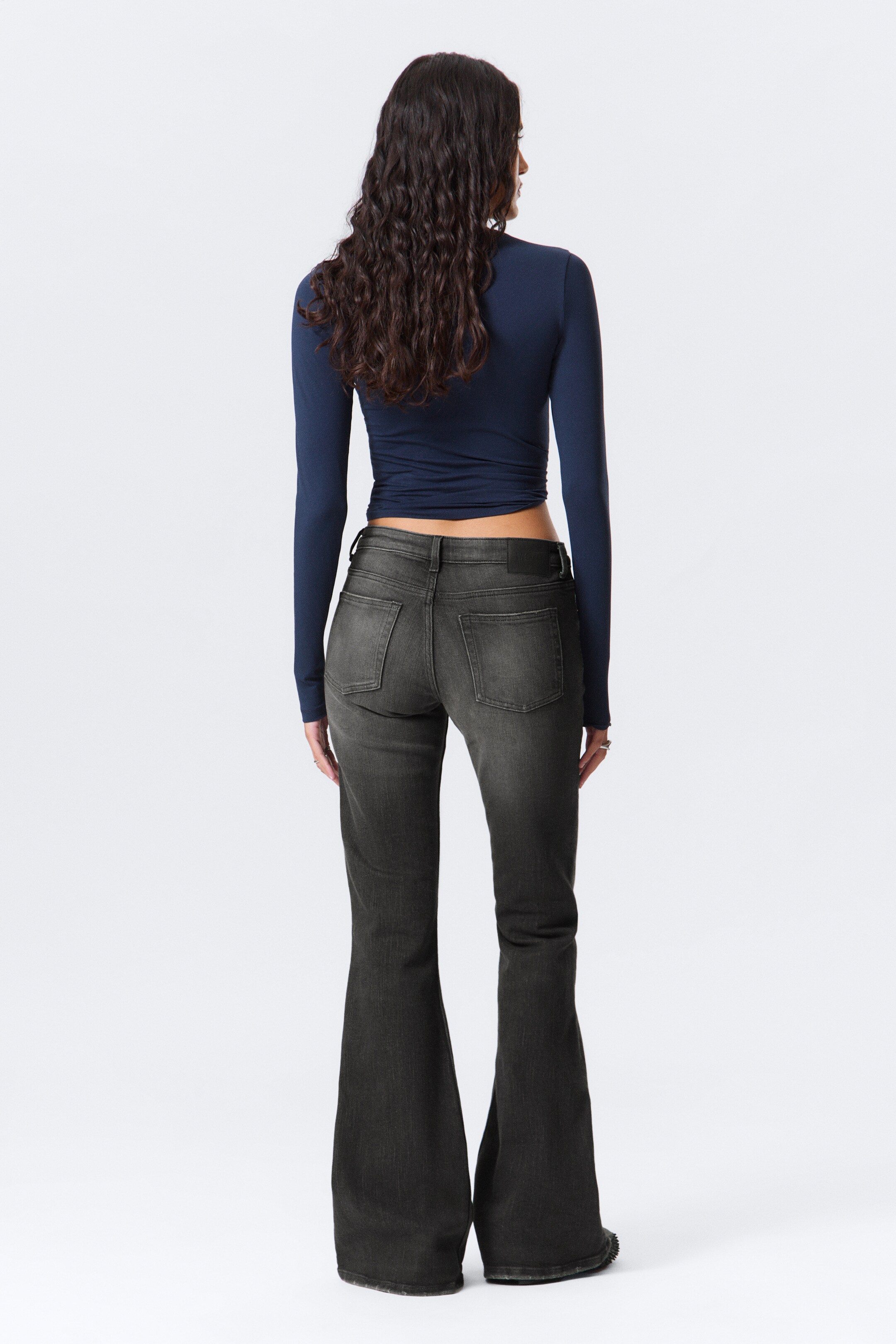 Grotere afbeelding bekijken: Donkergrijze uitlopende jeans worden gepresenteerd, bekeken van achteren. Ze hebben een aansluitend silhouet bij de heupen en dijen, met een uitgesproken flair vanaf de knieën, en bevatten twee achterzakken. Er wordt een donkerblauwe top met lange mouwen gedragen, waarbij de tailleband van de jeans zichtbaar is.