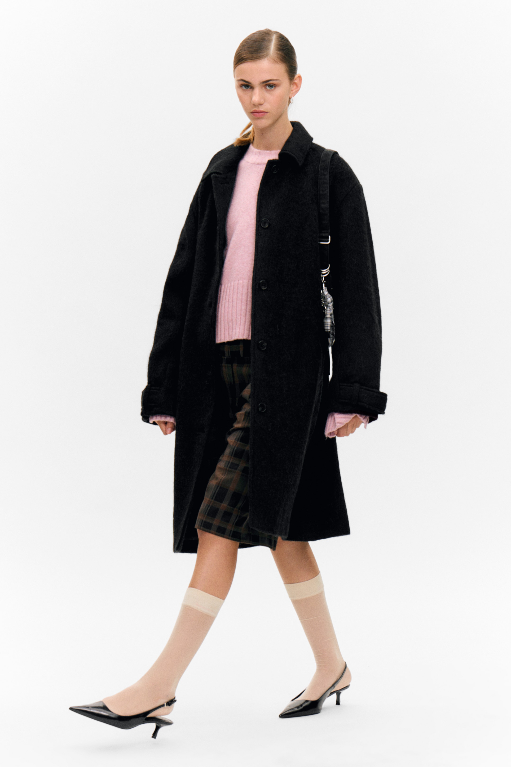 View larger image: A black, textured single-breasted coat is being worn from a slight front-side angle, featuring a classic collar and three visible front buttons. A pink sweater with ribbed cuffs is extending from the coat's sleeves, with plaid shorts visible underneath the coat.