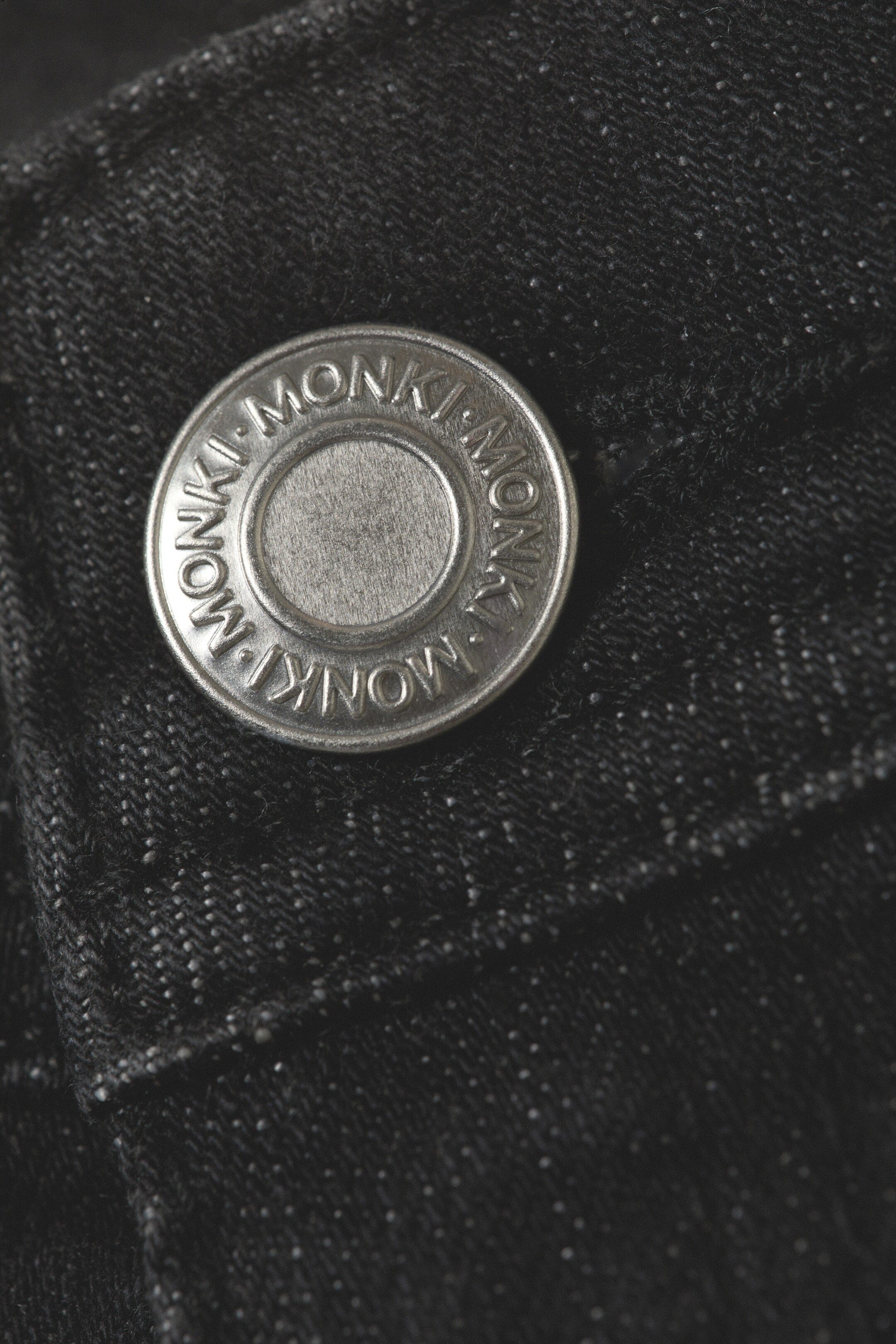 Agrandir l'image: Un bouton de couleur argent est attaché à un tissu en jean foncé. Le bouton est embossé avec 'MONKI' autour de son bord et présente un centre en métal brossé, tandis que le tissu en jean foncé montre un tissage visible à chevrons avec de subtiles taches blanches.