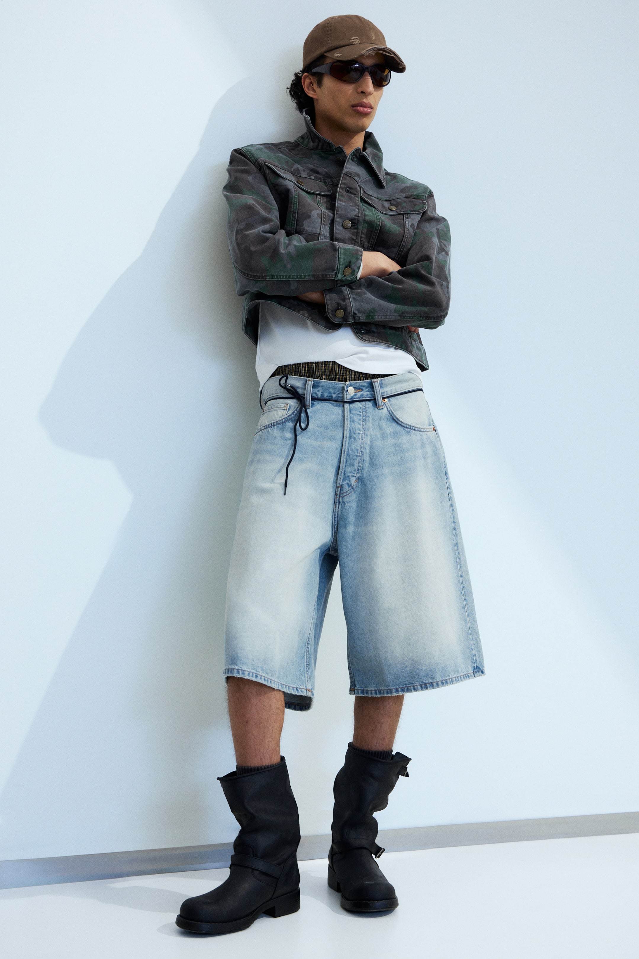 Agrandir l'image: Une tenue est présentée sous un angle légèrement frontal, comprenant une casquette de baseball marron vieilli, des lunettes de soleil foncées, et une veste en denim camouflage raccourcie avec des fermetures à boutons, superposée à un t-shirt blanc. L'ensemble inclut également un short en denim délavé clair à jambes larges descendant sous le genou, avec une taille à cordon de serrage, et des bottes noires mi-mollet avec des détails de boucles.