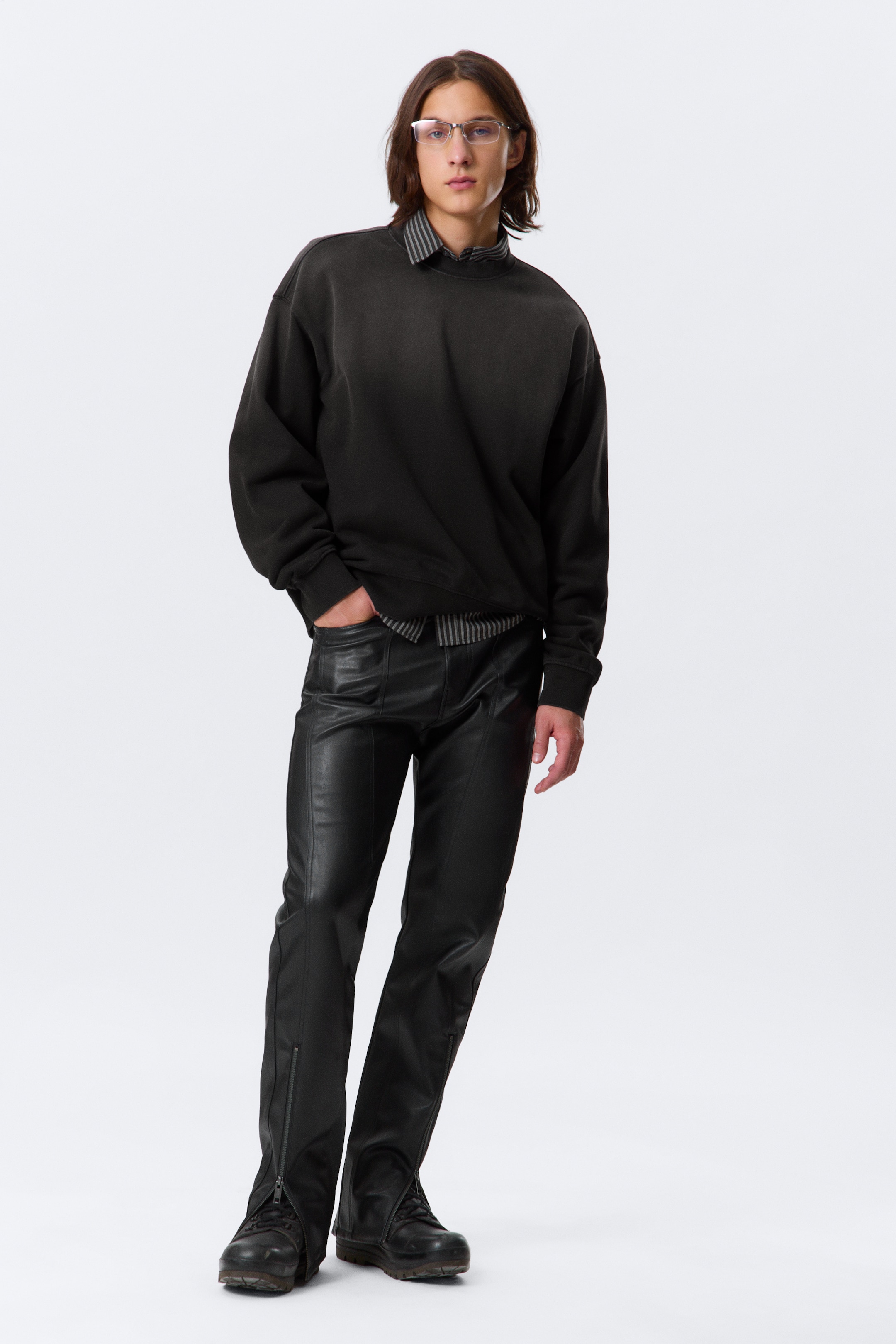 Visualizza immagine più grande: Una felpa oversize grigio scuro con scollo rotondo e un effetto sbiadito viene indossata, stratificata su una camicia a righe con colletto, e abbinata a pantaloni neri aderenti effetto pelle. I pantaloni presentano dettagli di cuciture visibili e cerniere alle caviglie, con stivali scuri visibili sotto.