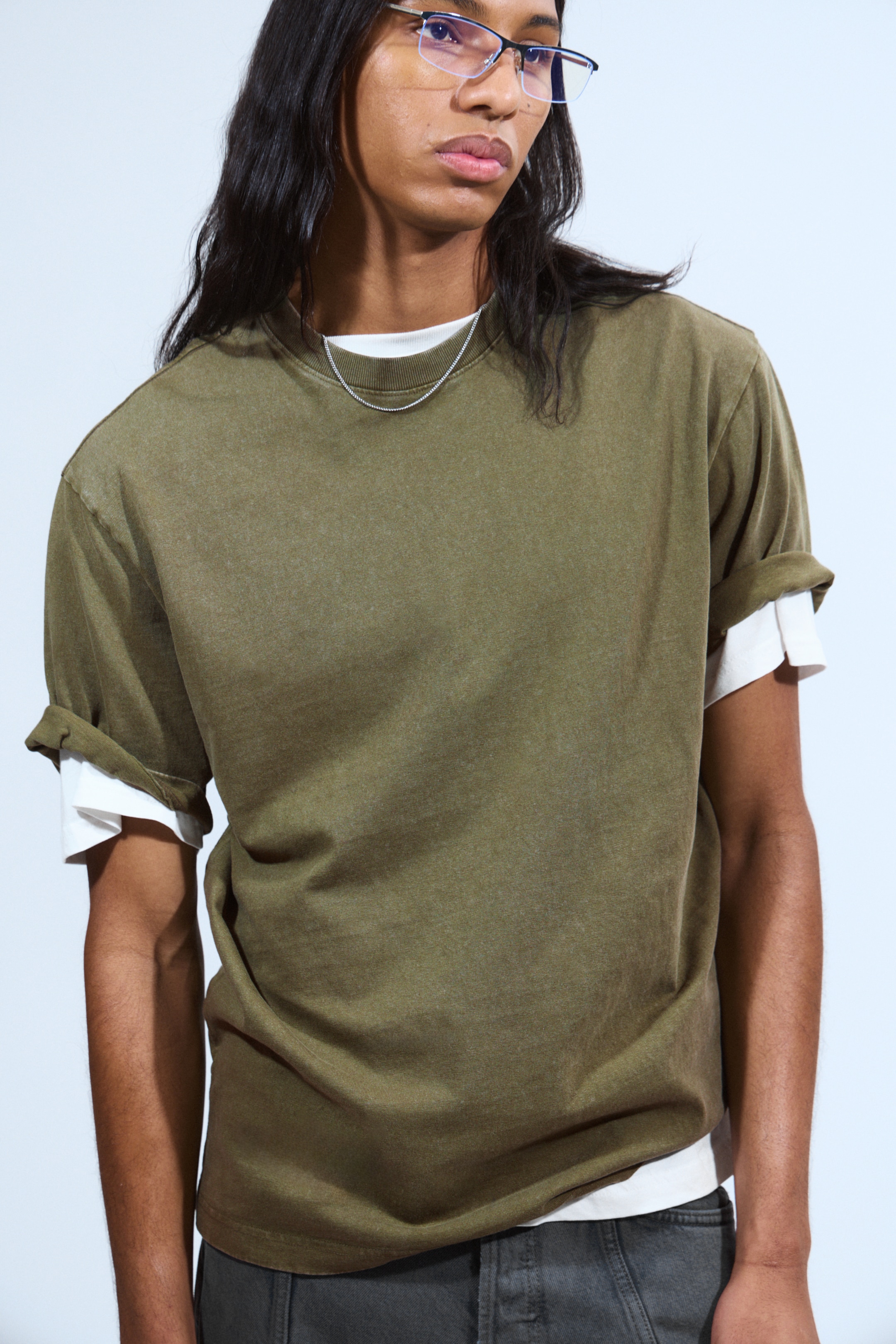 View larger image: An olive green T-shirt is being worn, seen from the front, slightly angled to the left. Its crew neck is showing, and its short sleeves are being rolled up, revealing a white long-sleeve shirt underneath. The T-shirt is displaying a washed, faded appearance.