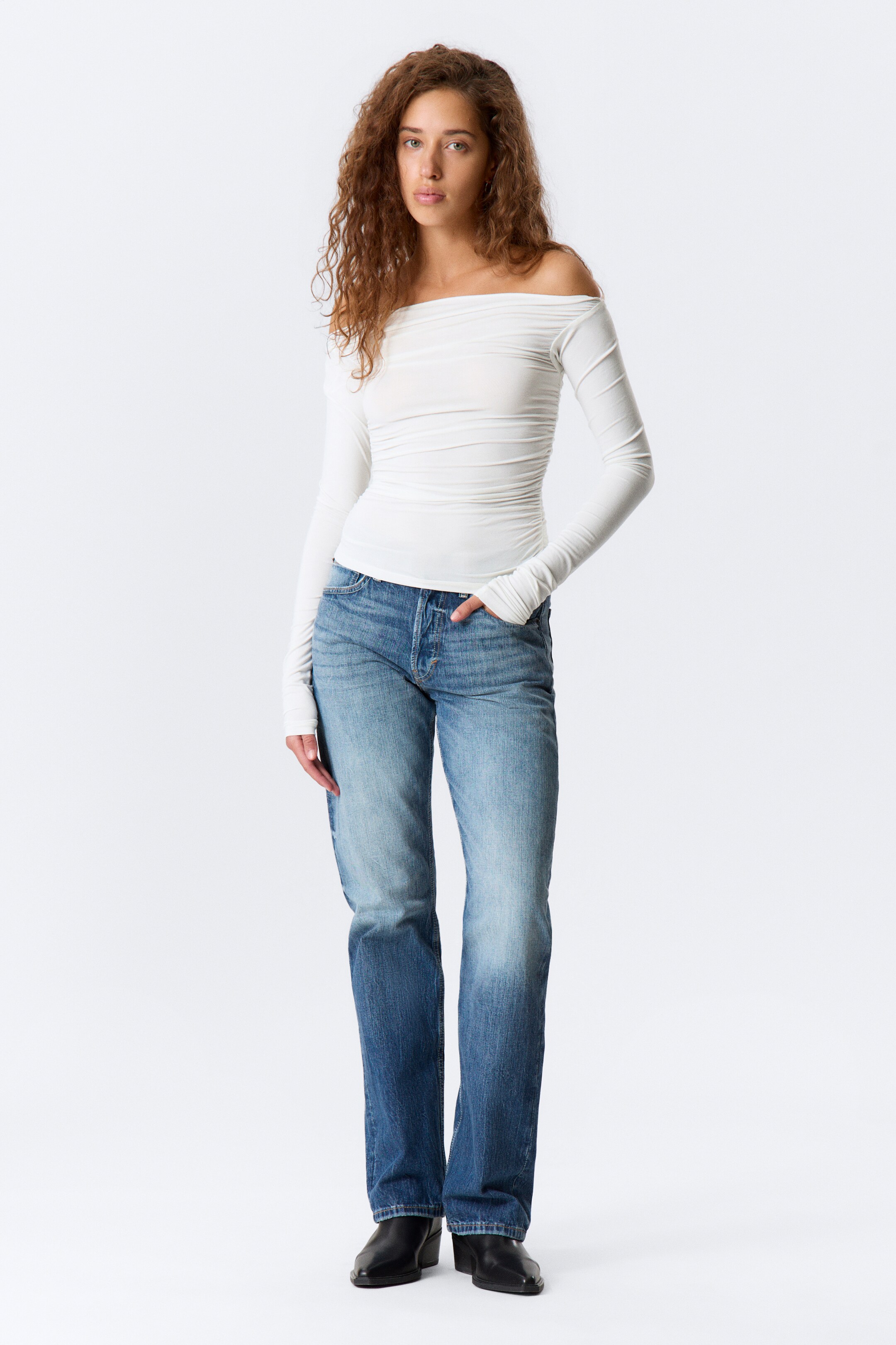 Agrandir l'image: Un haut blanc à manches longues et épaules dénudées, avec des détails froncés, est présenté de face. Il est porté avec un jean bleu à jambes droites, qui présente un effet délavé et une braguette à boutons apparente, et des bottes noires à bout pointu.