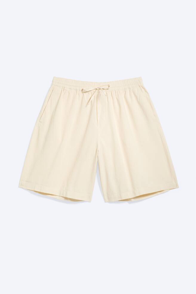 Short beige clair posés à plat et orientés vers l'avant, dotés d'une taille élastiquée avec un cordon de serrage noué et des poches latérales visibles. - 1