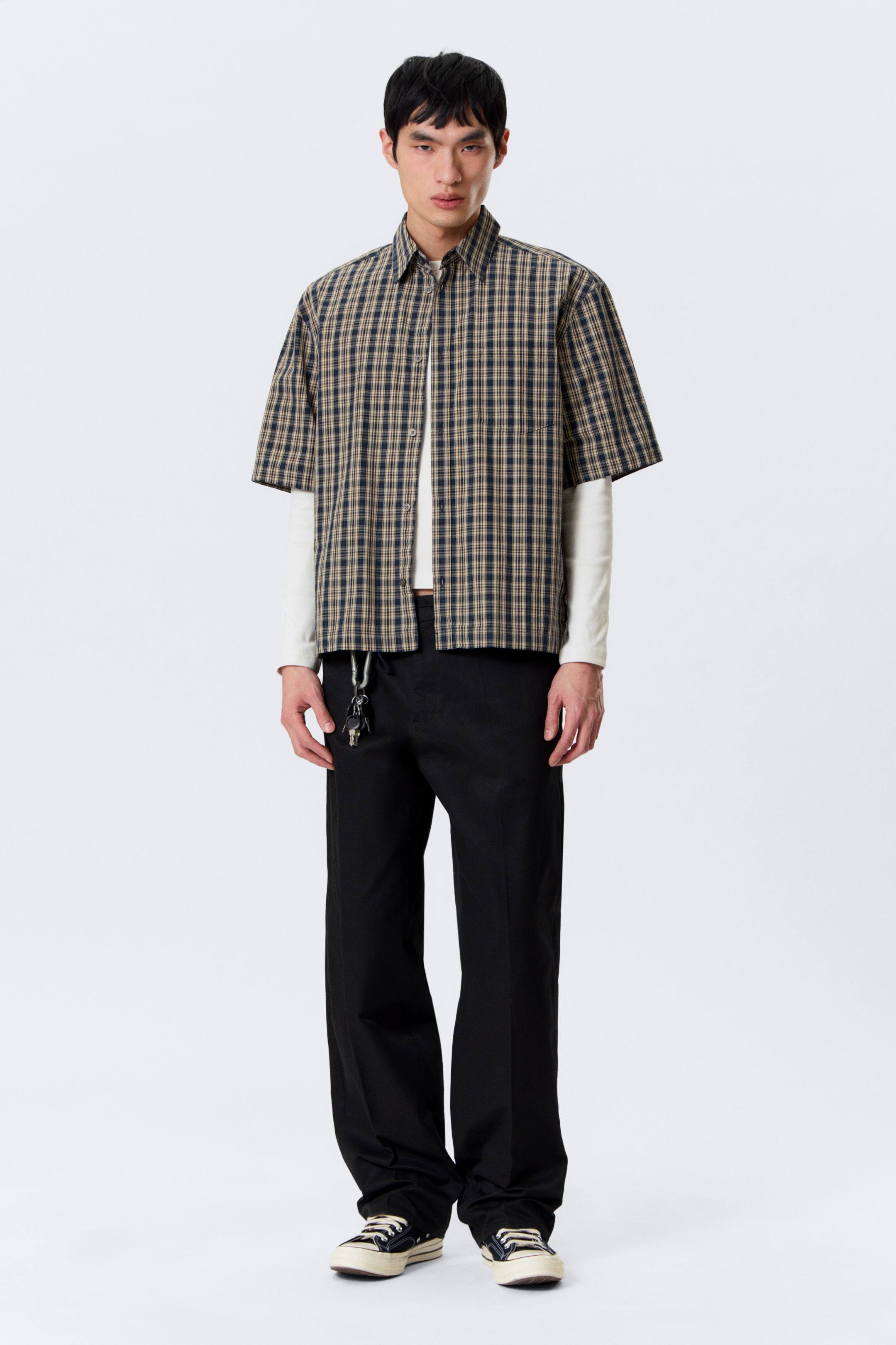 Ingrandisci l'immagine: Una camicia a maniche corte a quadri presenta un motivo a scacchi blu scuro e beige, con un classico colletto a punta, una parte anteriore parzialmente sbottonata e una tasca applicata sul petto sinistro. È indossata sopra una maglia bianca a maniche lunghe.