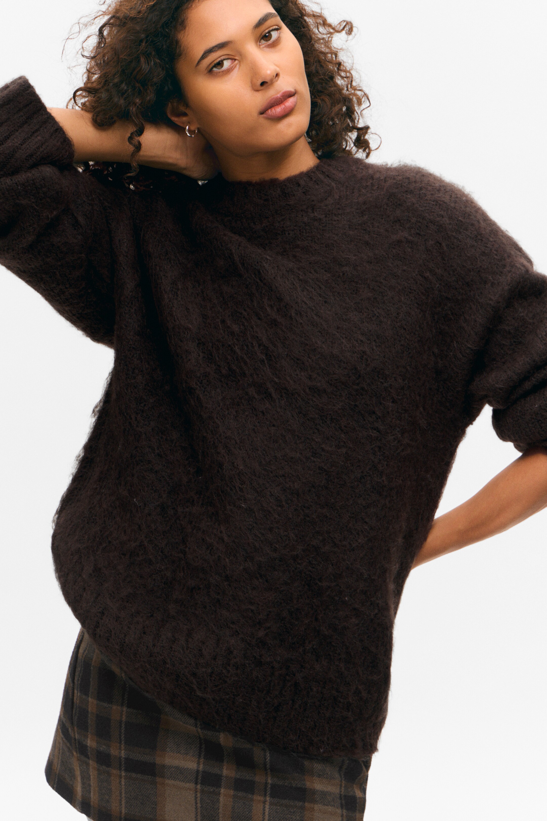 Ingrandisci l'immagine: Un maglione a girocollo in maglia marrone scuro e peloso viene indossato in una vestibilità oversize, leggermente angolato a sinistra, con maniche lunghe e polsini a coste, stratificato sopra una gonna a quadri.