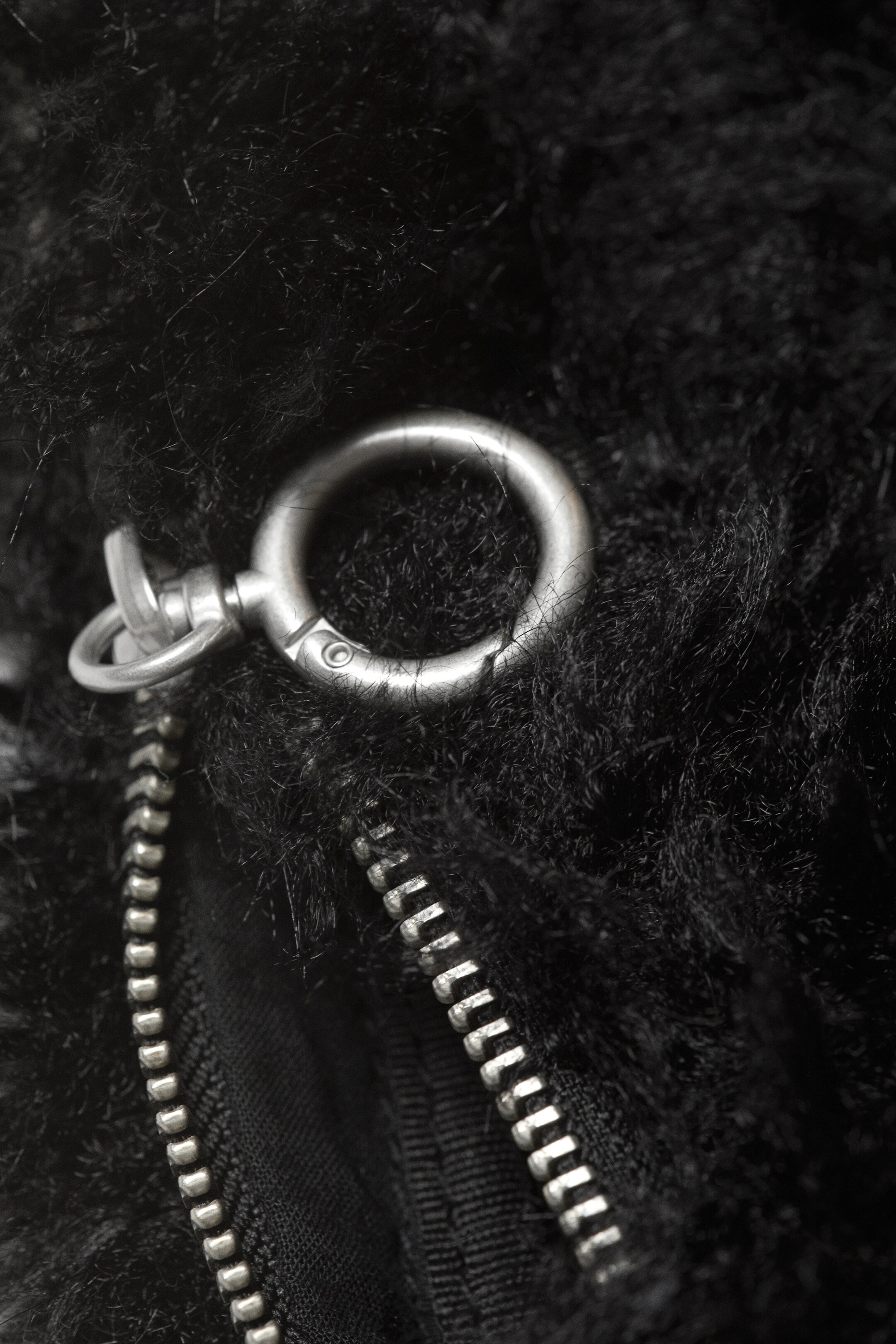 Größeres Bild ansehen: Ein schwarzes, langfloriges Kunstfellmaterial ist mit einem großen, mattsilbernen O-Ring mit Drehverschluss ausgestattet, der über einem teilweise geöffneten silberfarbenen Metallreißverschluss positioniert ist.