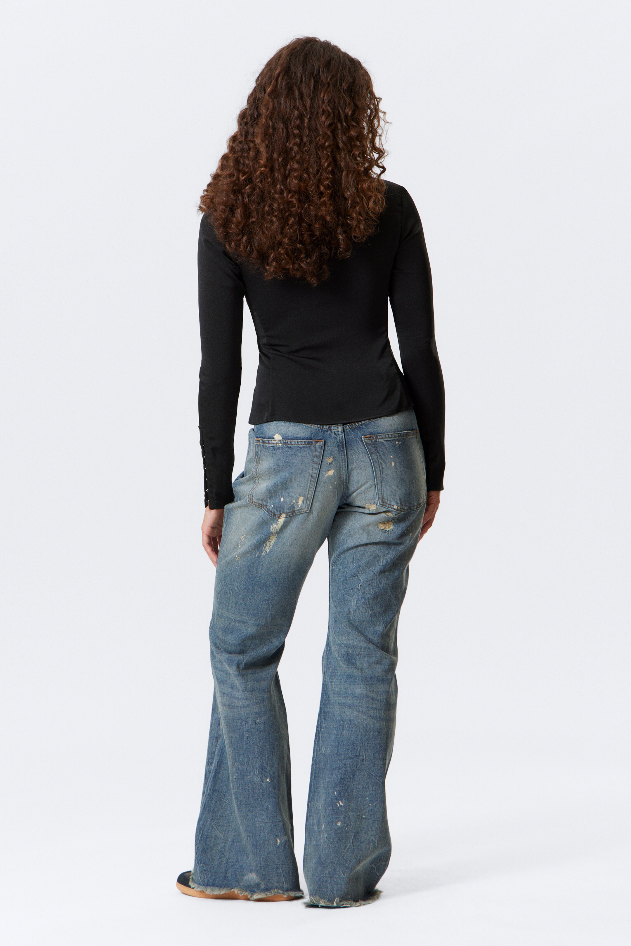 View larger image: A black long-sleeved top is being worn from the back, displaying a fitted silhouette with button details along the forearms. It is being paired with light blue, distressed denim jeans, which are featuring a loose, flared cut, visible rips, paint splatters, and frayed hems.