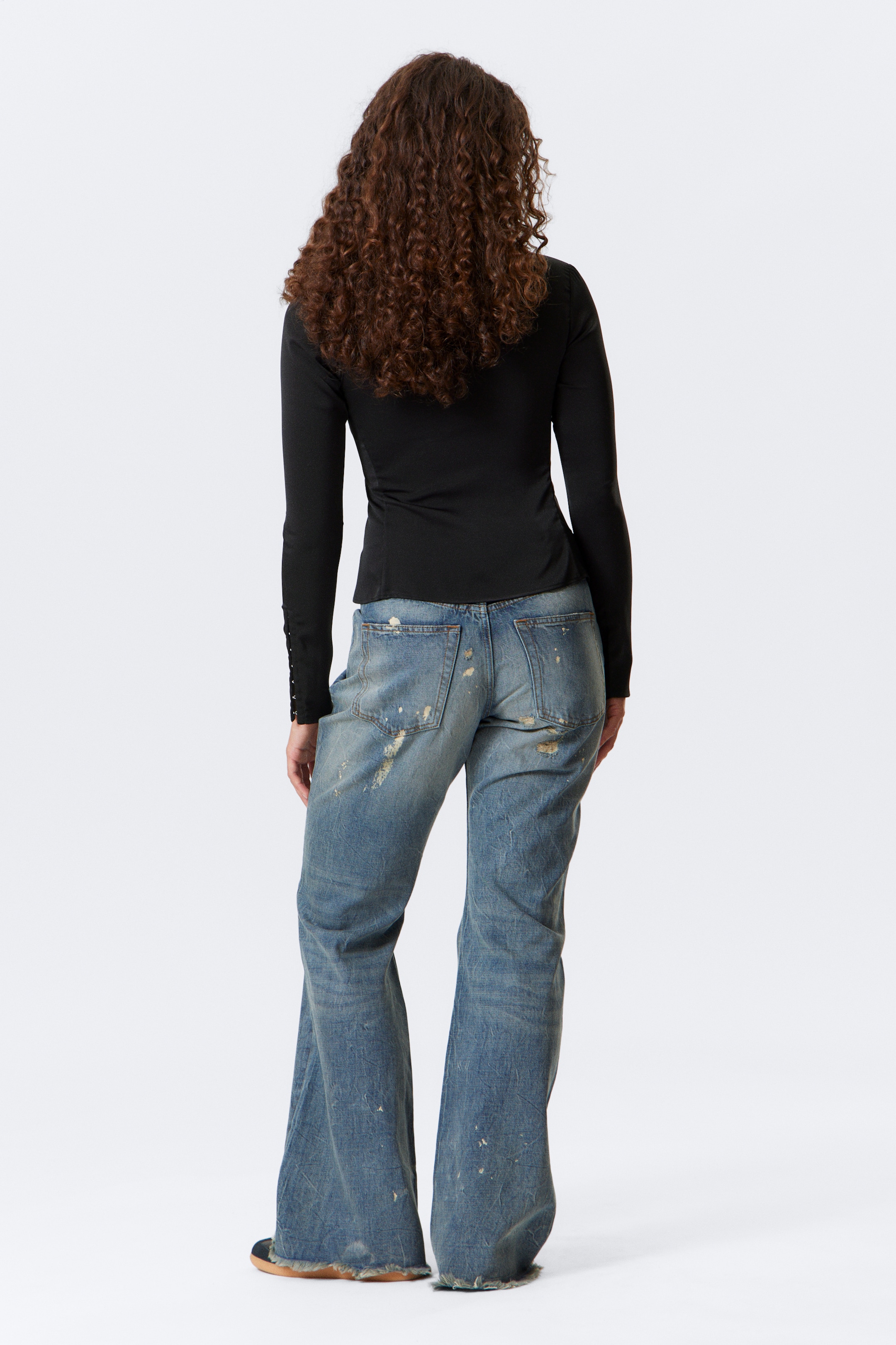 Se større billede: En sort langærmet top bæres fra ryggen, og viser en tætsiddende silhuet med knapdetaljer langs underarmene. Den er parret med lyseblå, slidte denimjeans, som har et løst, svajet snit, synlige revner, malerpletter og flossede kanter.