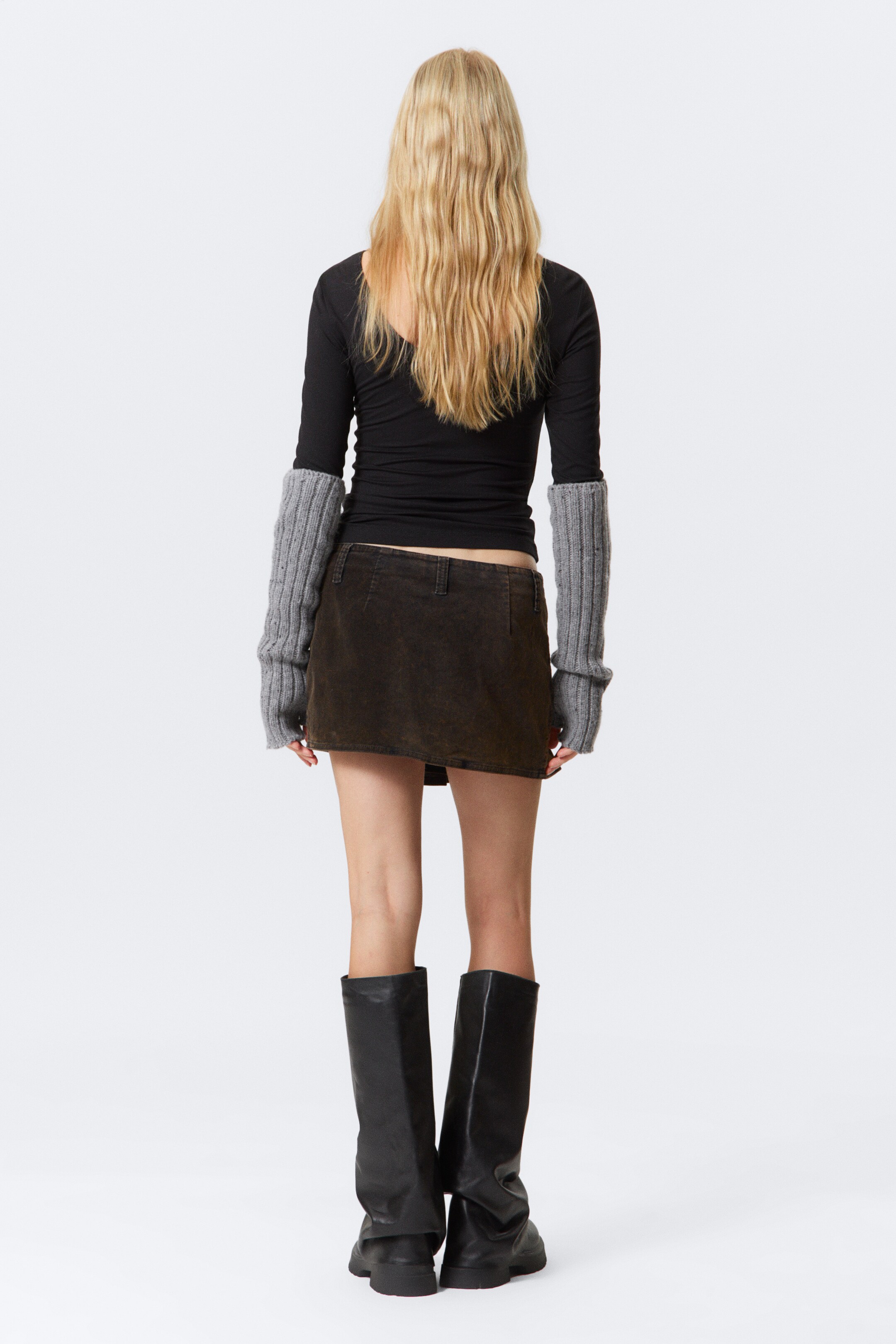 Grotere afbeelding bekijken: Een bruine mini-rok met textuur wordt van de achterkant gepresenteerd, met riemlussen en een rechte zoomlijn. Het wordt gestyled met een zwarte top met lange mouwen, grijze geribbelde gebreide armwarmers en zwarte slouchy kniehoge laarzen.