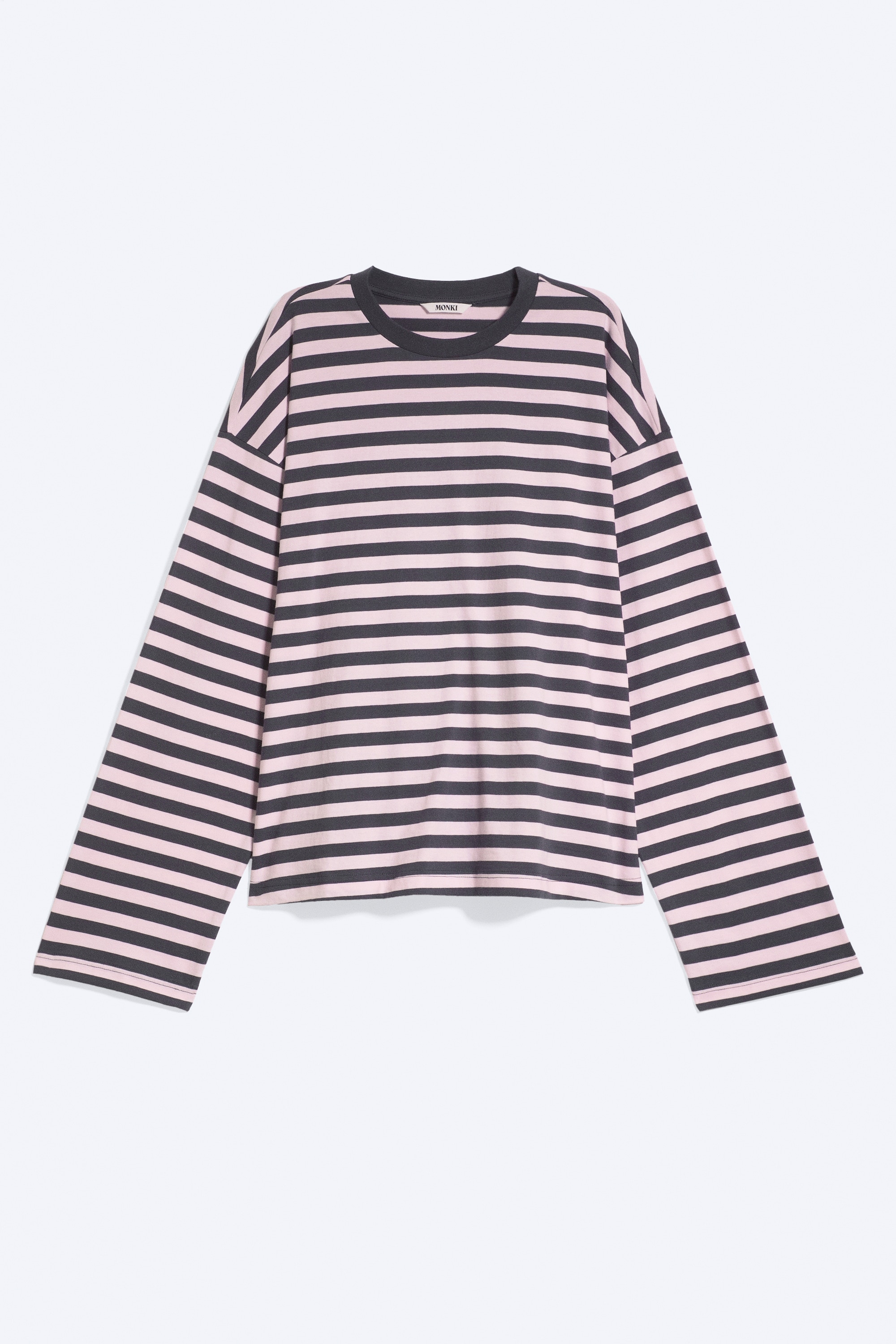 Ένα μακρυμάνικο T-shirt με άνετη εφαρμογή και ροζ-μαύρες οριζόντιες ρίγες φοριέται από το μοντέλο. Το T-shirt διαθέτει κλασική στρογγυλή λαιμόκοψη και ελαφρώς πεσμένους ώμους, και συνδυάζεται με ανοιχτόχρωμο μπλε τζιν, καθώς το μοντέλο στέκεται με σταυρωμένα τα χέρια.