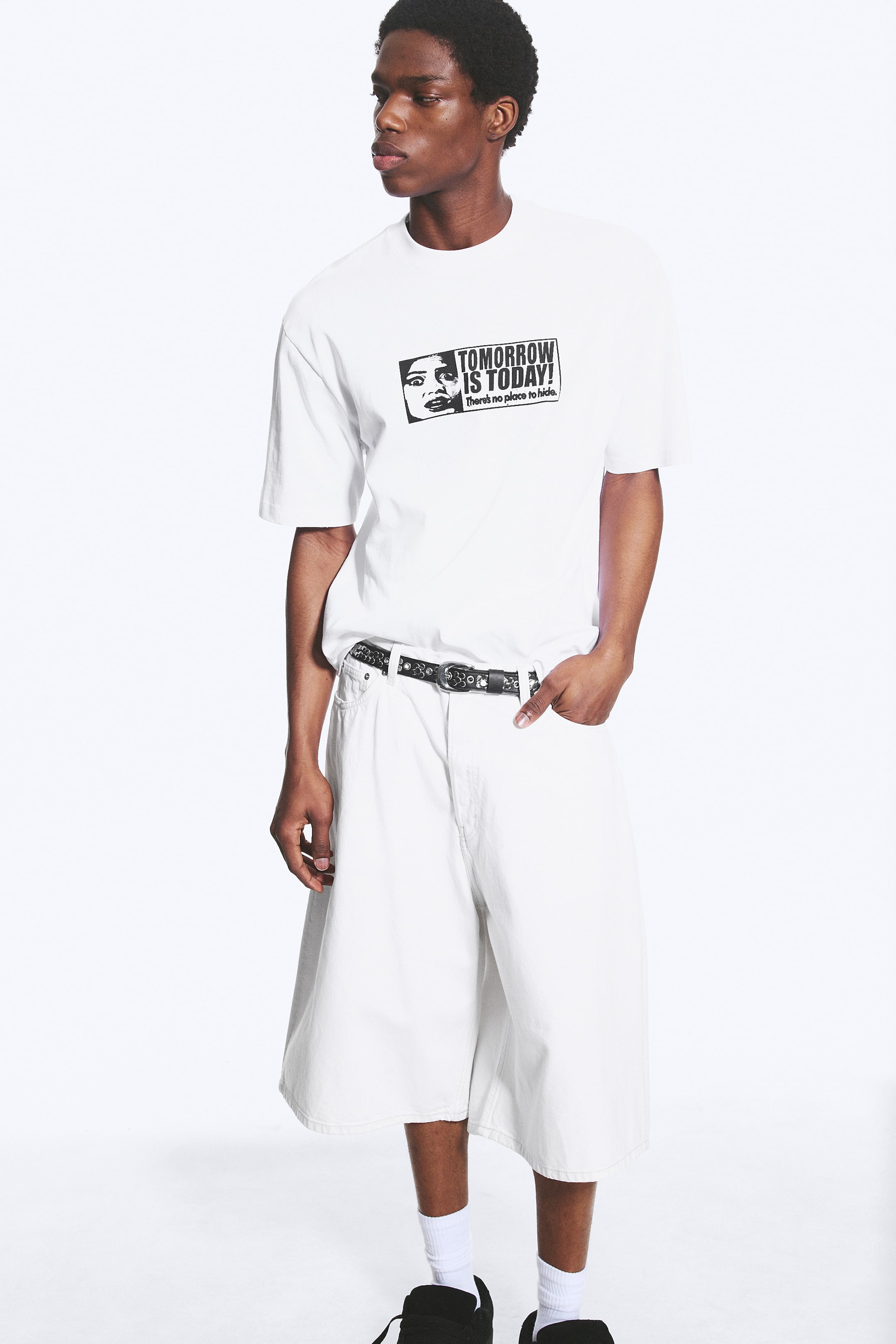 Ver imagen más grande: Una camiseta blanca de manga corta presenta un gráfico negro en el pecho, que muestra el rostro de una mujer y el texto "¡EL MAÑANA ES HOY! No hay lugar para esconderse". Esta camiseta se combina con pantalones cortos blancos de pierna ancha y un cinturón negro con una hebilla de tono plateado y un patrón en relieve. El conjunto se muestra ligeramente orientado hacia la derecha.
