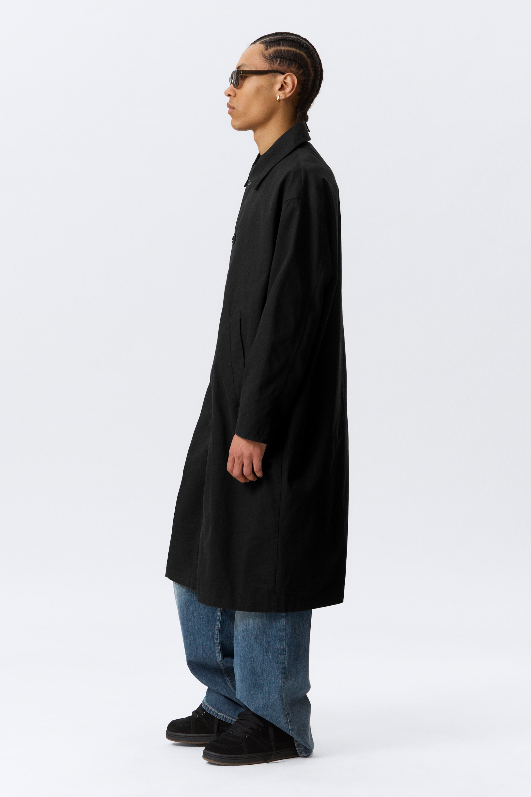 Agrandir l'image: Un manteau noir présente une coupe décontractée et droite vue de profil. Il est doté d'un col classique en pointe, de manches longues et de poches dans les coutures latérales, s'étendant juste en dessous des genoux sur un jean bleu.