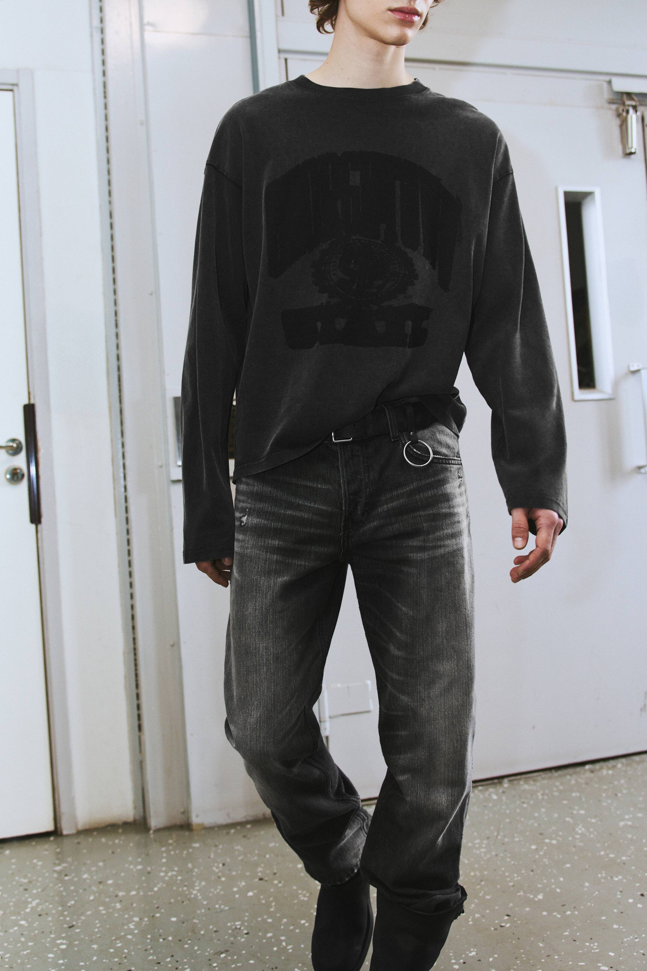 Agrandir l'image: Un T-shirt à manches longues à la coupe boxy, en gris foncé délavé, doté d’un col rond et d’un imprimé graphique délavé sur la poitrine, est porté par le mannequin. Le T-shirt est stylisé avec l’ourlet avant partiellement rentré dans un jean gris foncé délavé.
