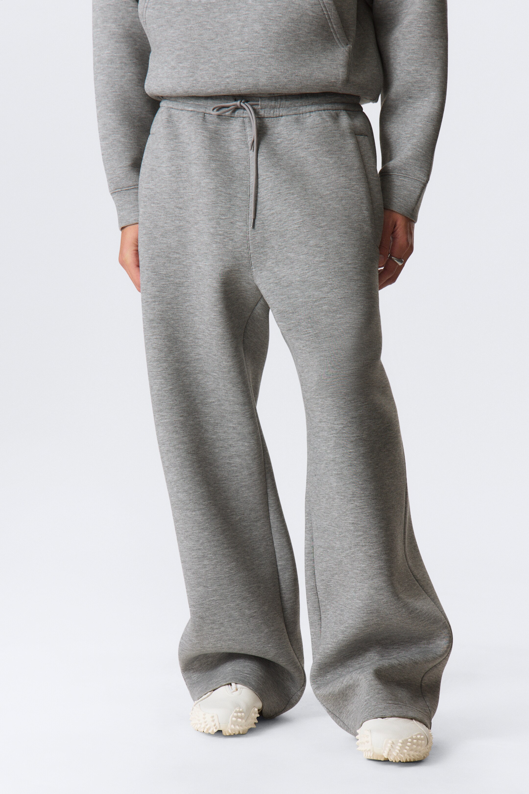 Agrandir l'image: Les pantalons de survêtement gris à jambes larges sont dotés d'une ceinture élastiquée avec un cordon de serrage visible à l'avant. Ils sont coupés avec une coupe décontractée, fabriqués à partir d'un tissu gris doux, et tombent largement aux chevilles, reposant sur des chaussures blanches.
