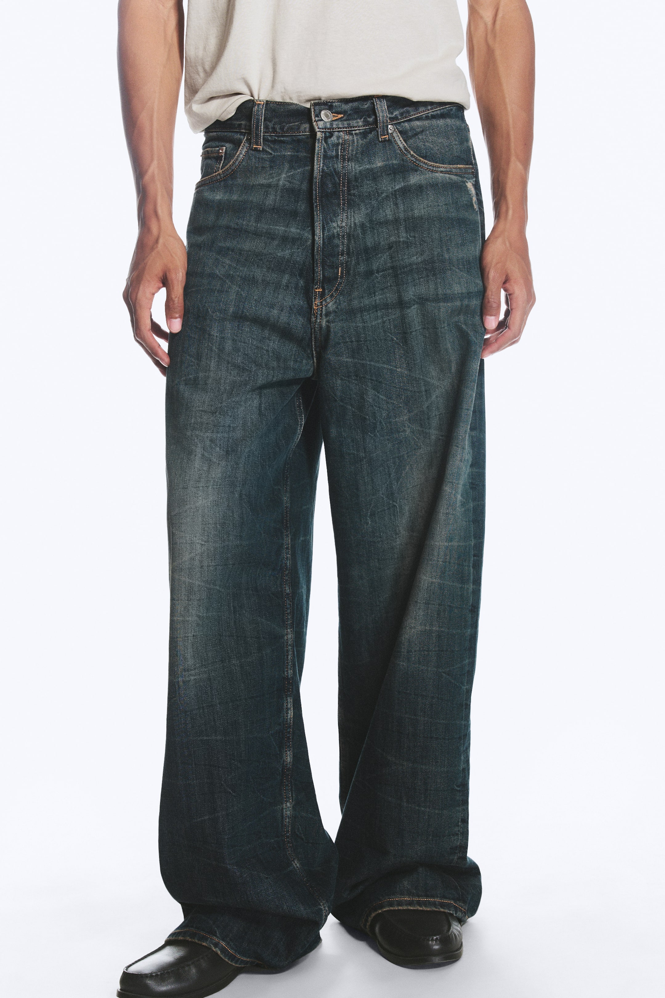 Grotere afbeelding bekijken: Het model draagt een paar loose-fit jeans met middelhoge taille in een donkere, vervaagde wassing. De jeans hebben een baggy pijpsilhouet dat over donkere schoenen valt, een klassiek vijfzakkenontwerp en een knoopsluiting in de taille, waar een lichtgekleurde T-shirt ingestopt is.