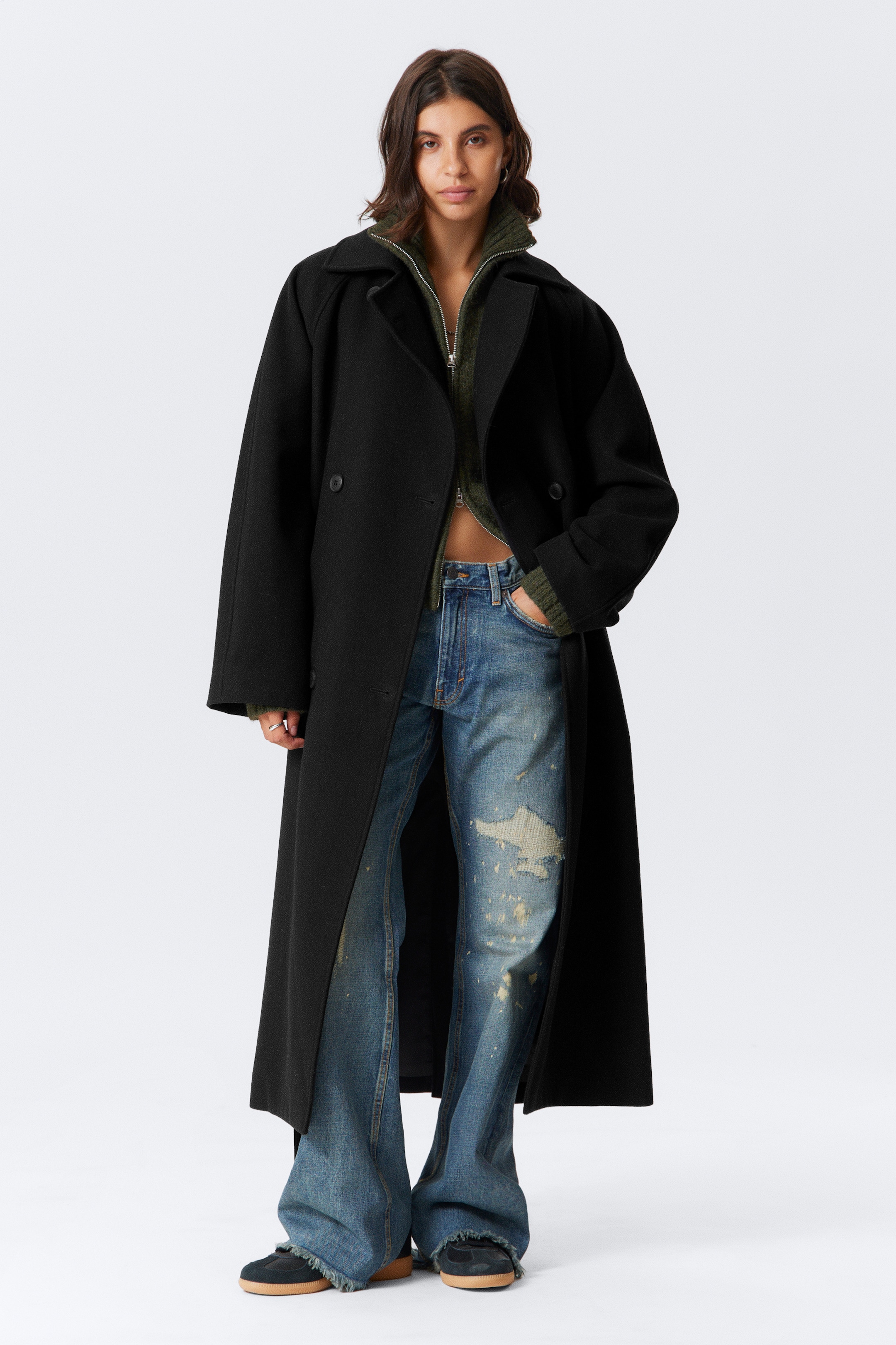 Visualizza immagine più grande: Un lungo cappotto nero doppiopetto, caratterizzato da ampie revers a lancia, maniche raglan e una silhouette oversize, è indossato aperto dalla modella, la cui mano è appoggiata in una tasca a filetto, sopra un top verde e jeans strappati.