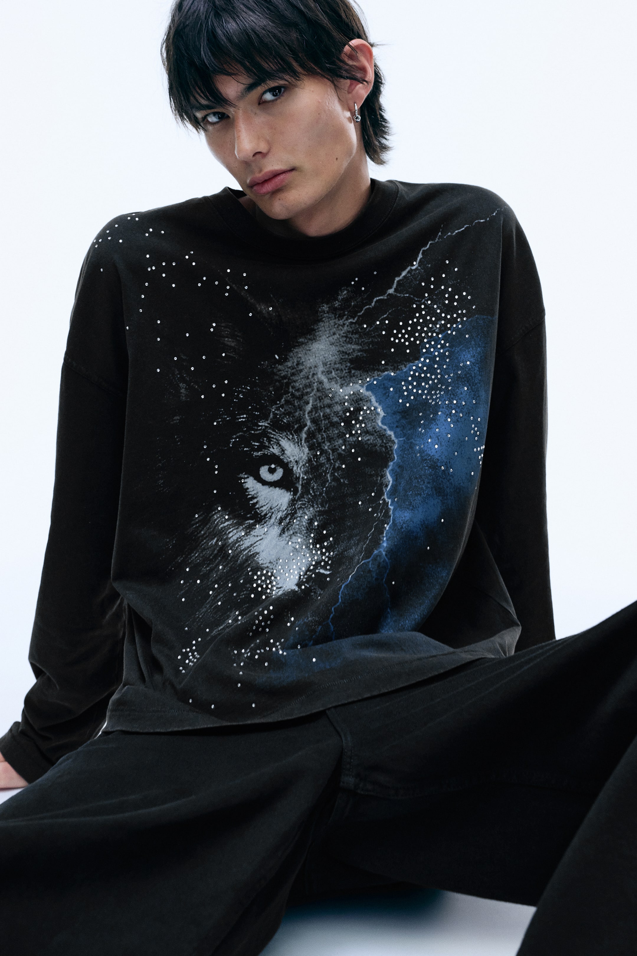 Visualizza immagine più grande: Una t-shirt nera oversize a maniche lunghe, con una grafica del volto di un lupo, fulmini e nebulose blu con dettagli effetto strass, è indossata dalla modella. La t-shirt è abbinata a pantaloni neri.