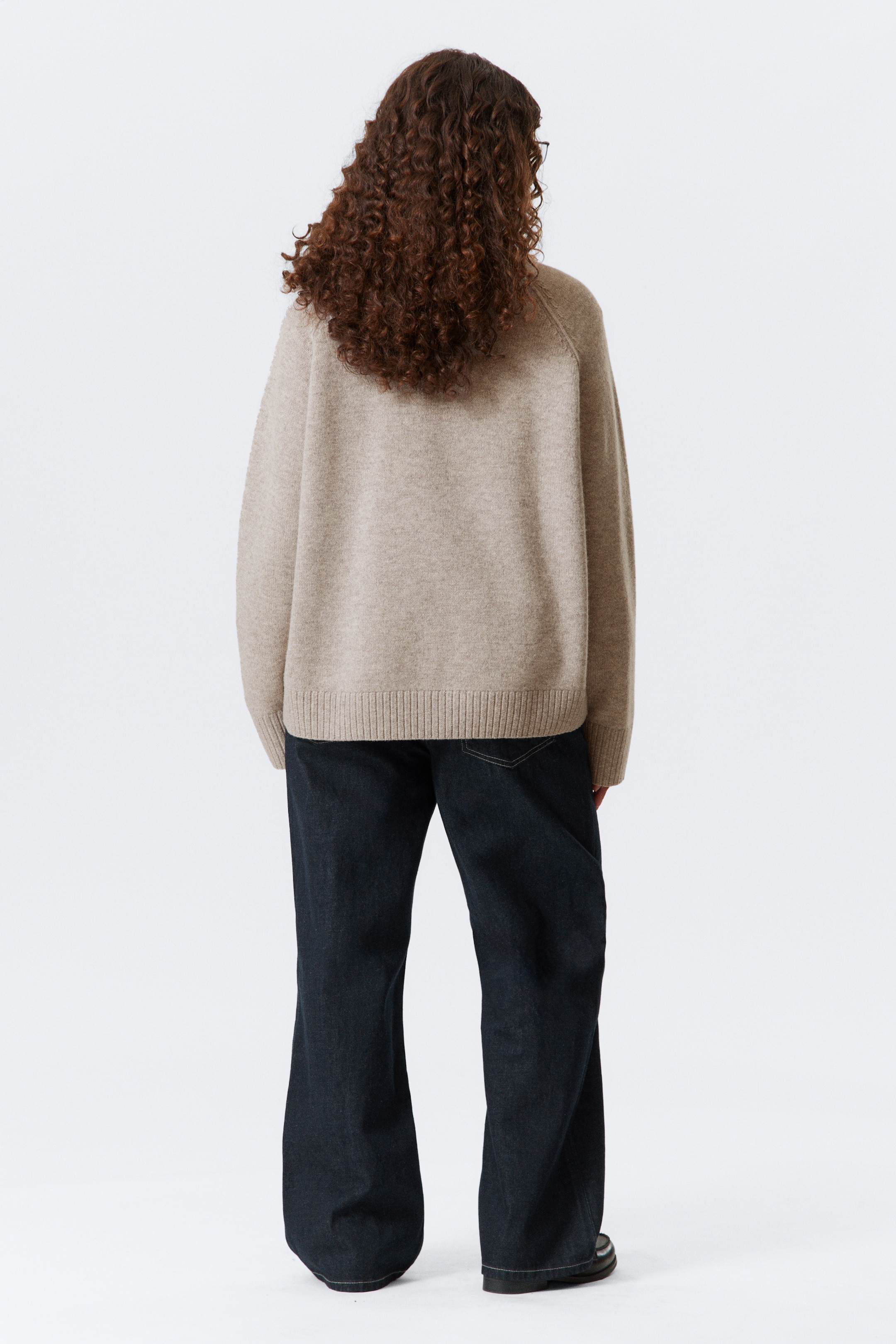 Größeres Bild ansehen: Ein beigefarbener Strickpullover wird von hinten gezeigt, mit einer lockeren Passform, geripptem Saum und gerippten Bündchen. Er wird mit dunkel gewaschenen, weit geschnittenen Jeans getragen.