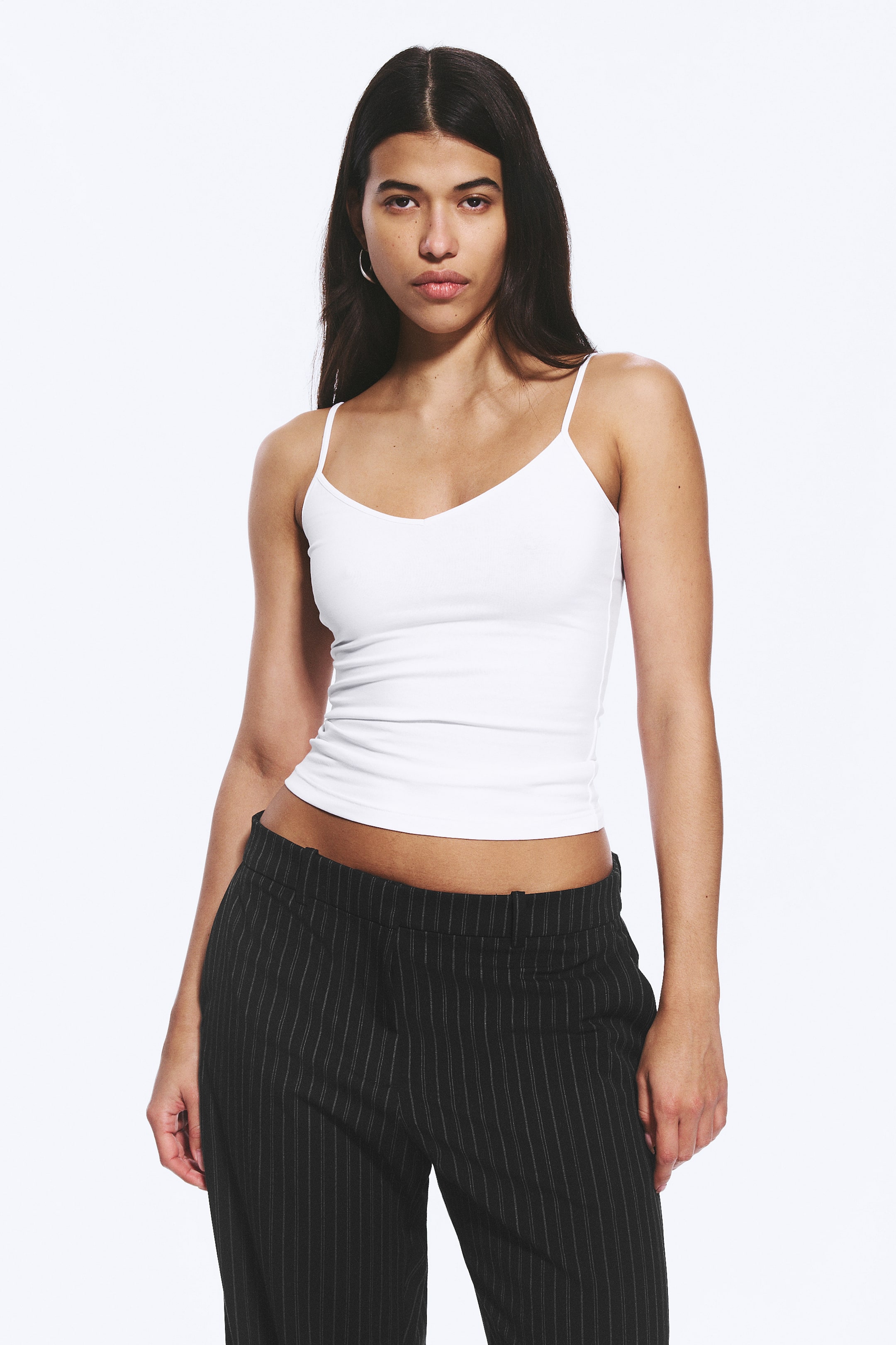Ver imagen más grande: Un camisole blanco acanalado con finos tirantes tipo espagueti y un escote en V, presenta una silueta ajustada y ligeramente recortada que termina por encima de la cintura de unos pantalones negros de rayas diplomáticas.