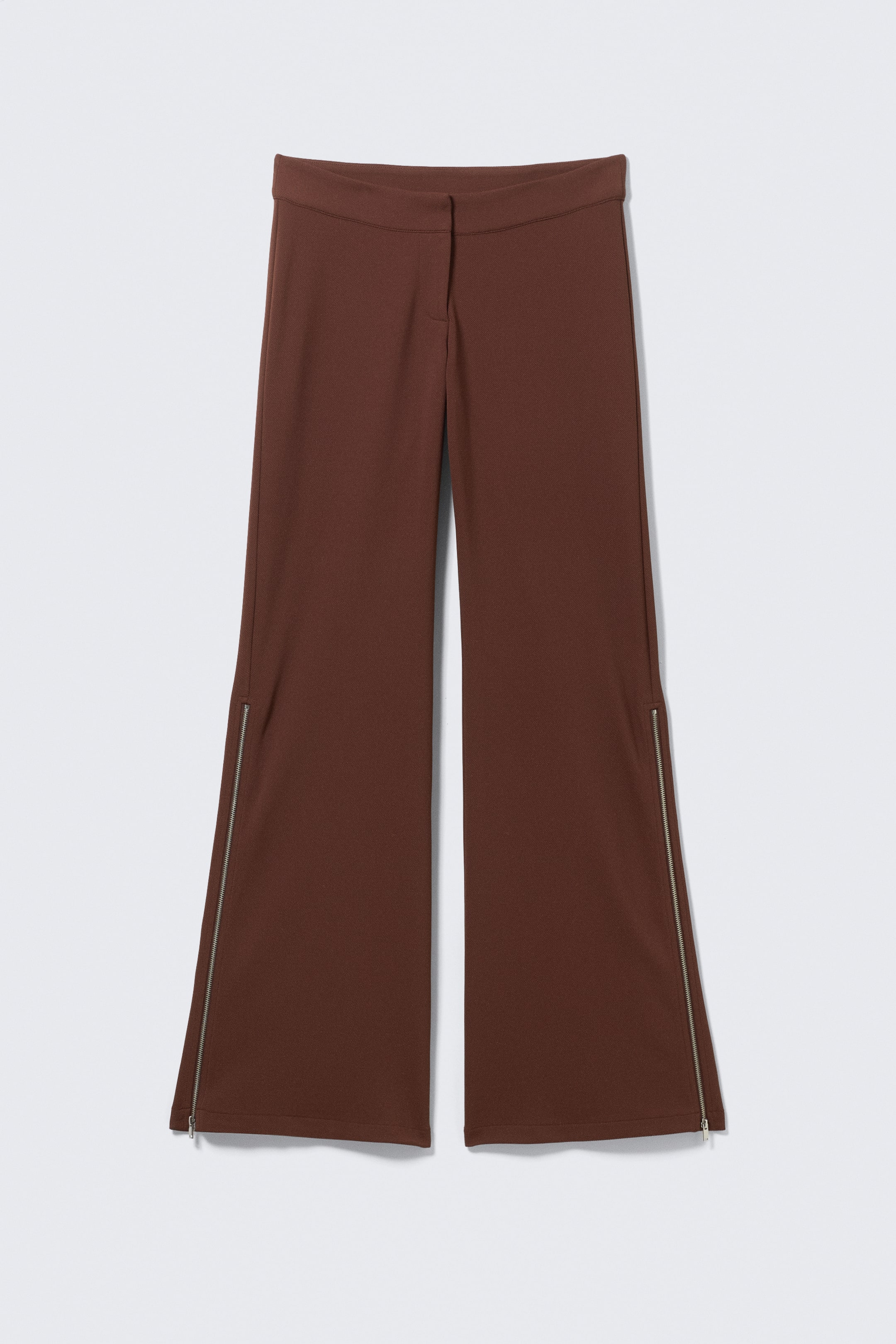 Größeres Bild ansehen: Die braunen Hosen präsentieren eine Vorderansicht und zeichnen sich durch einen flachen Bund und ein ausgestelltes Bein-Design aus. Silberfarbene Reißverschlüsse verlaufen entlang der äußeren Naht jedes Beins, von unterhalb des Knies bis zum Saum, und sind am unteren Ende teilweise geöffnet.