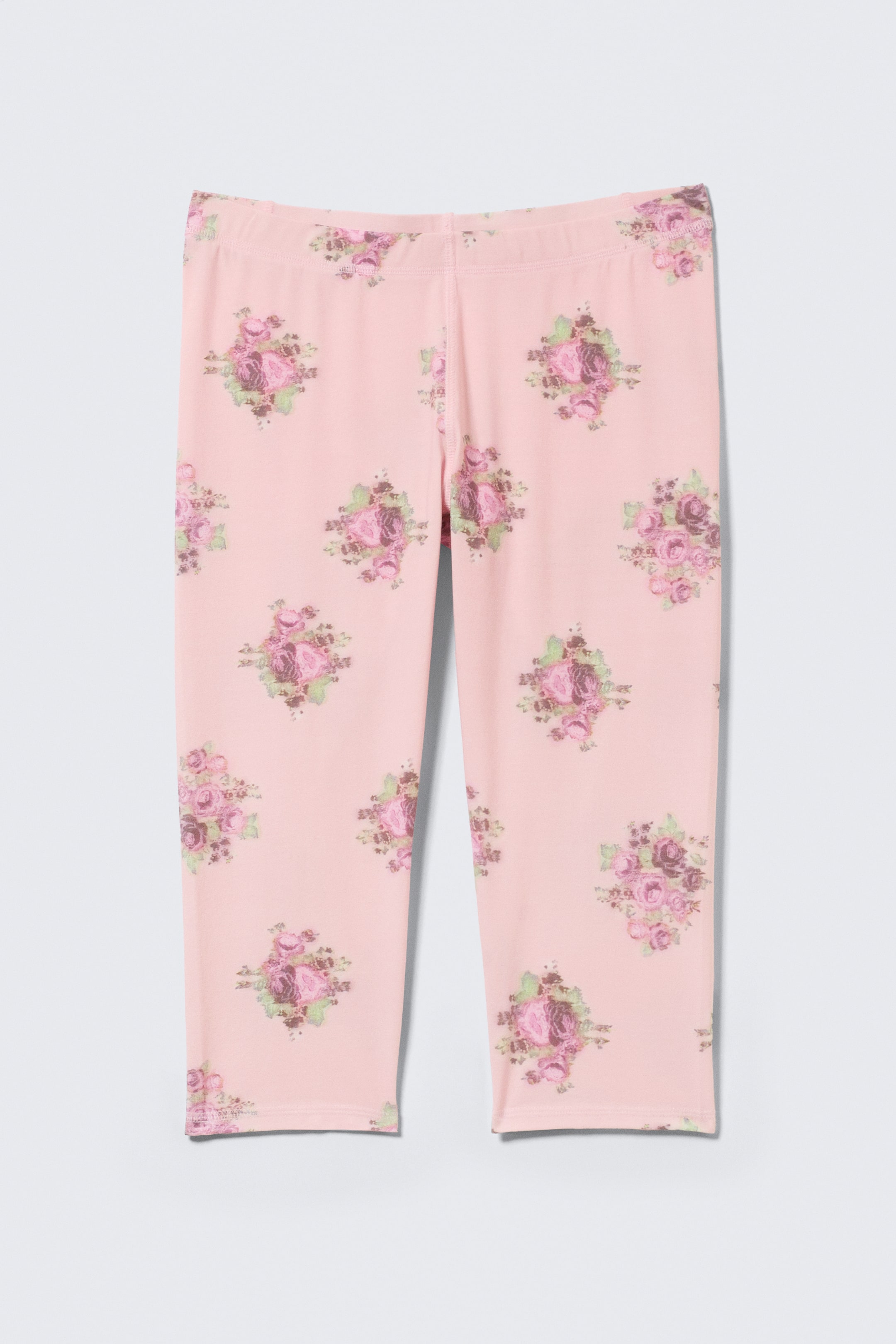 Ver imagen más grande: Un par de leggings capri se muestra desde el frente, apareciendo en un color rosa claro con un patrón floral de rosas rosas y moradas y hojas verdes. Los leggings presentan un largo tres cuartos, una cintura elástica de altura media y costuras visibles a lo largo de las piernas internas.