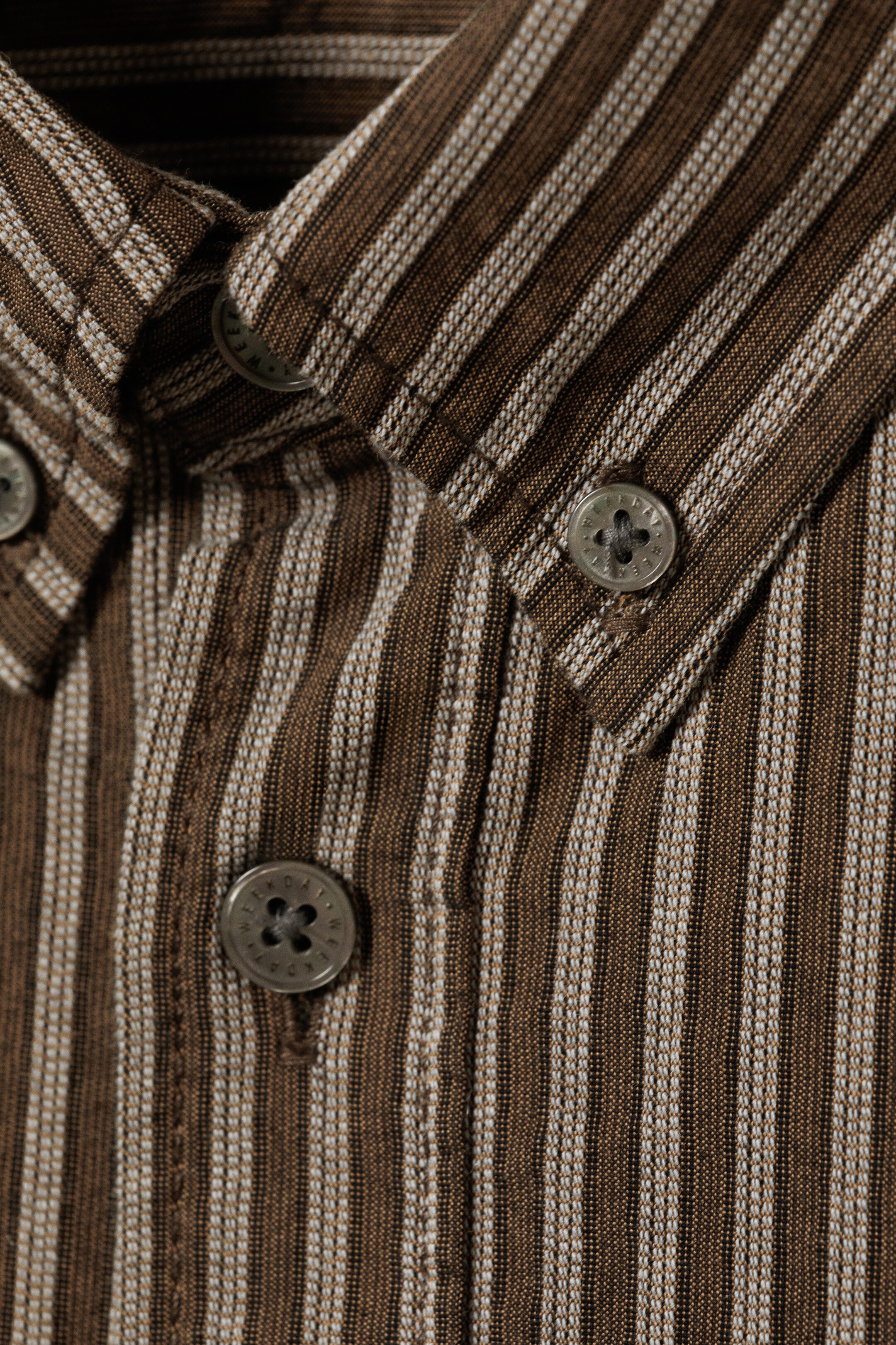 Größeres Bild anzeigen: Ein gestreiftes Hemd zeigt braune und cremeweiße vertikale Streifen über seinen Stoff. Es verfügt über einen Button-Down-Kragen mit einem sichtbaren Knopf an der Spitze, und dunkle, runde Knöpfe erscheinen auf der vorderen Knopfleiste.